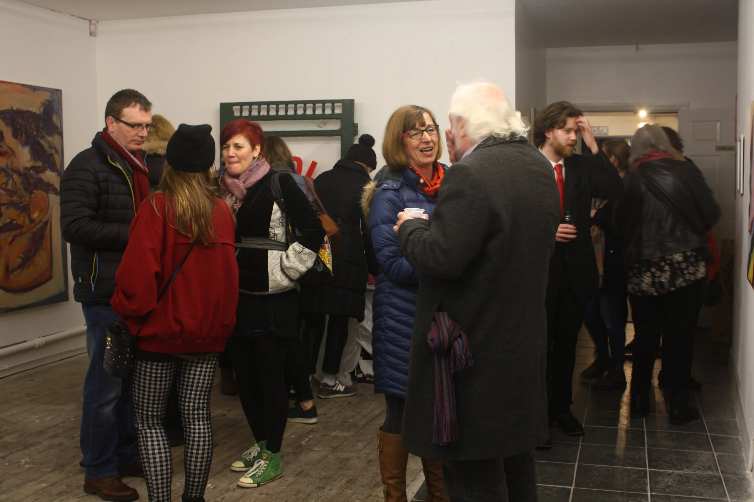People gathered and socializing at an art gallery exhibition, with artwork hanging on the white walls.