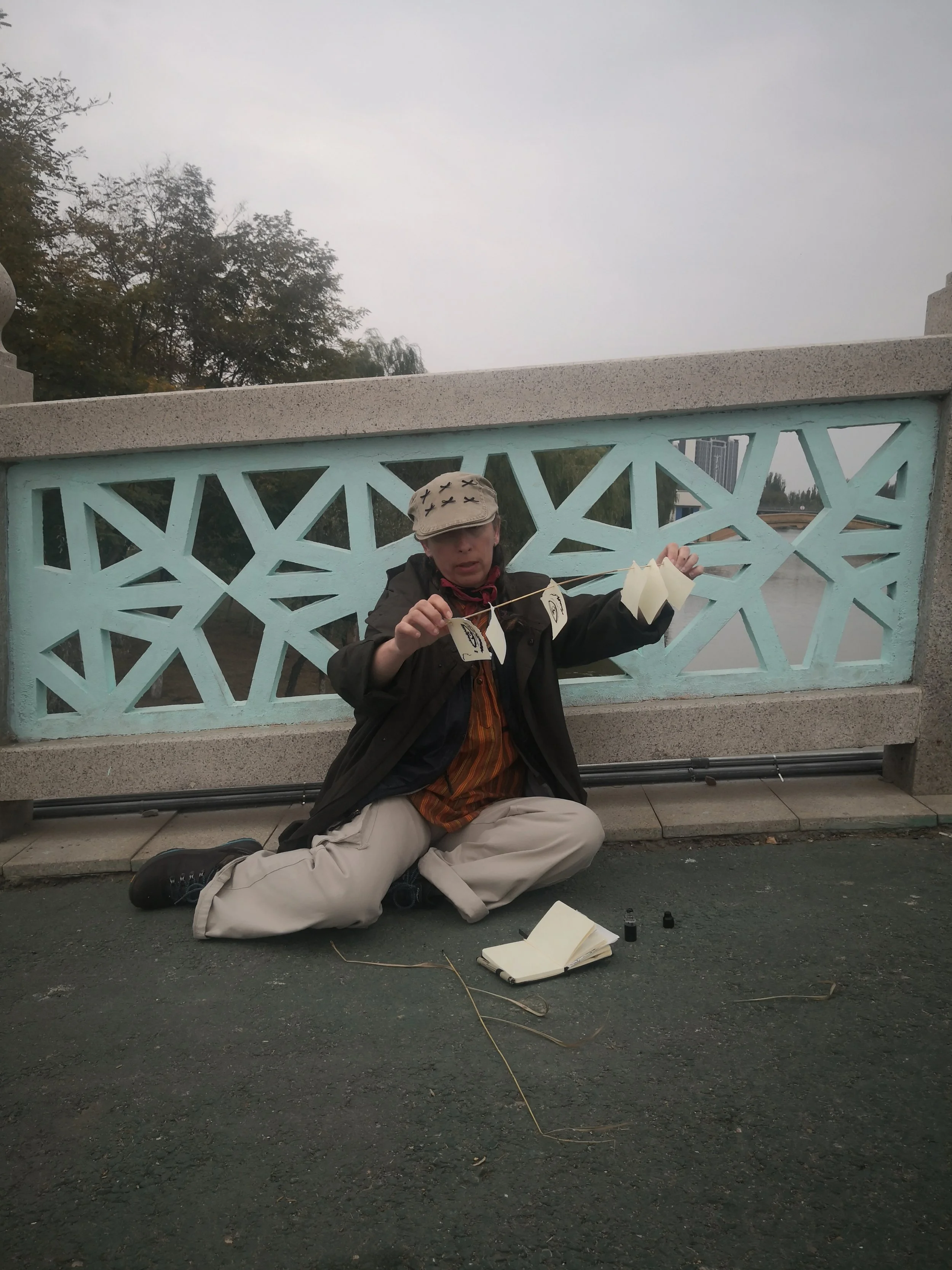 Person sitting on the ground in front of a decorative fence, holding a paper lantern. The person is wearing a beige hat with X marks, a dark jacket, and beige pants, with an open notebook and small bottles on the ground.