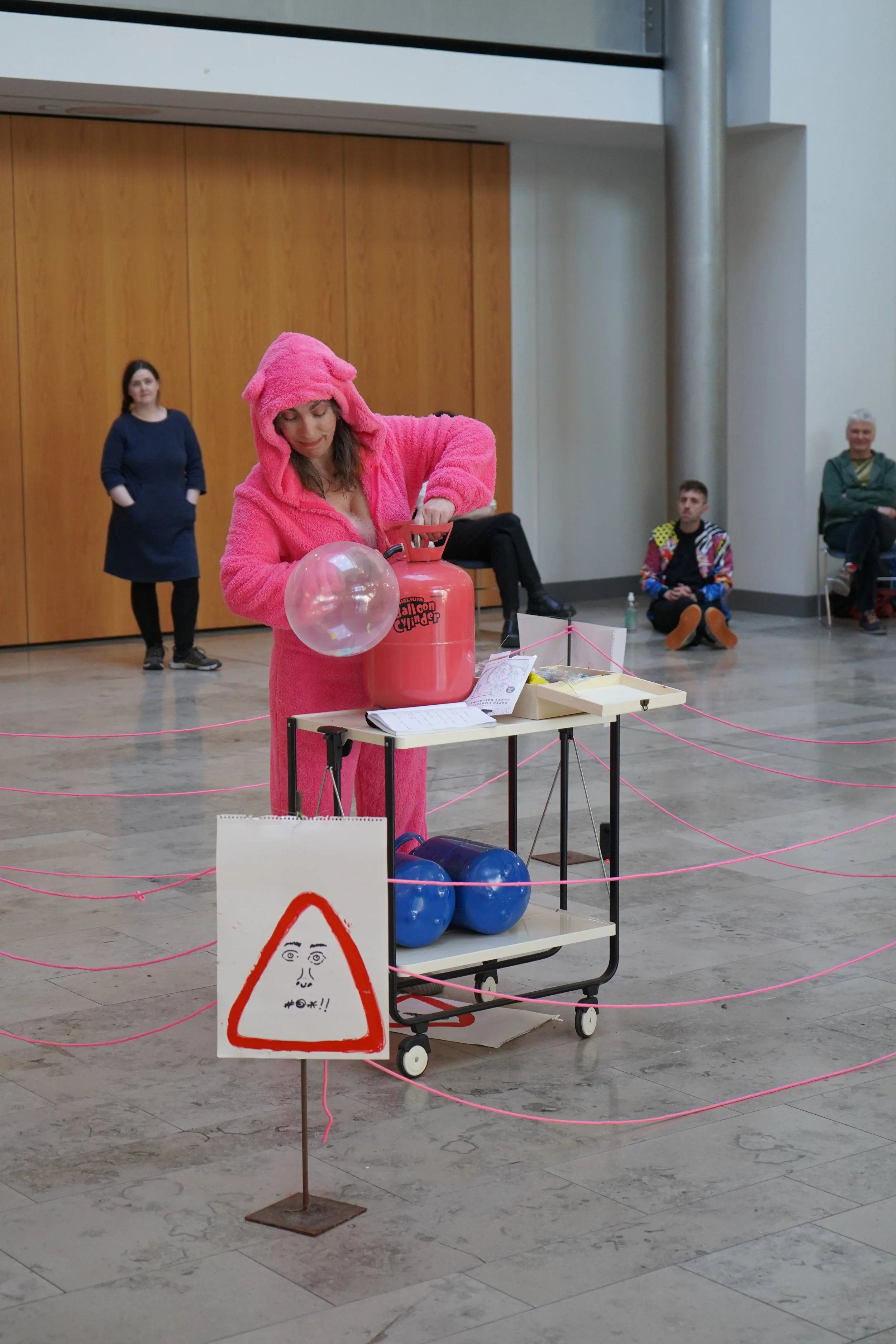 A woman in a pink bunny costume inflates a clear balloon with a fire extinguisher labeled "Balloon Cylinder" in an indoor open space. There are three people watching in the background, a woman in a navy dress, a young man in colorful clothing, and an