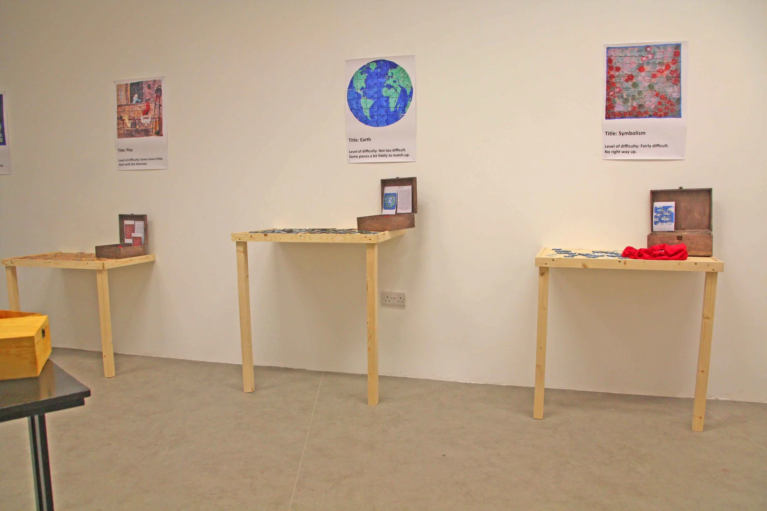 An indoor exhibition with three framed posters on white wall and three wooden tables with puzzle pieces and open boxes, likely part of an educational or puzzle activity display.
