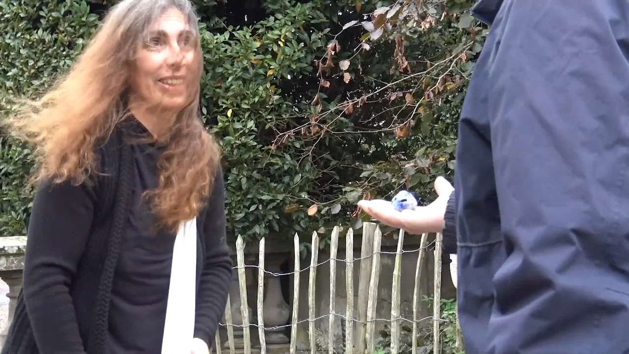 A woman with long, wavy hair smiling and facing a person in a blue jacket who is holding a small blue and white ceramic ball in their hand. They are outdoors near a wooden fence and lush green bushes.