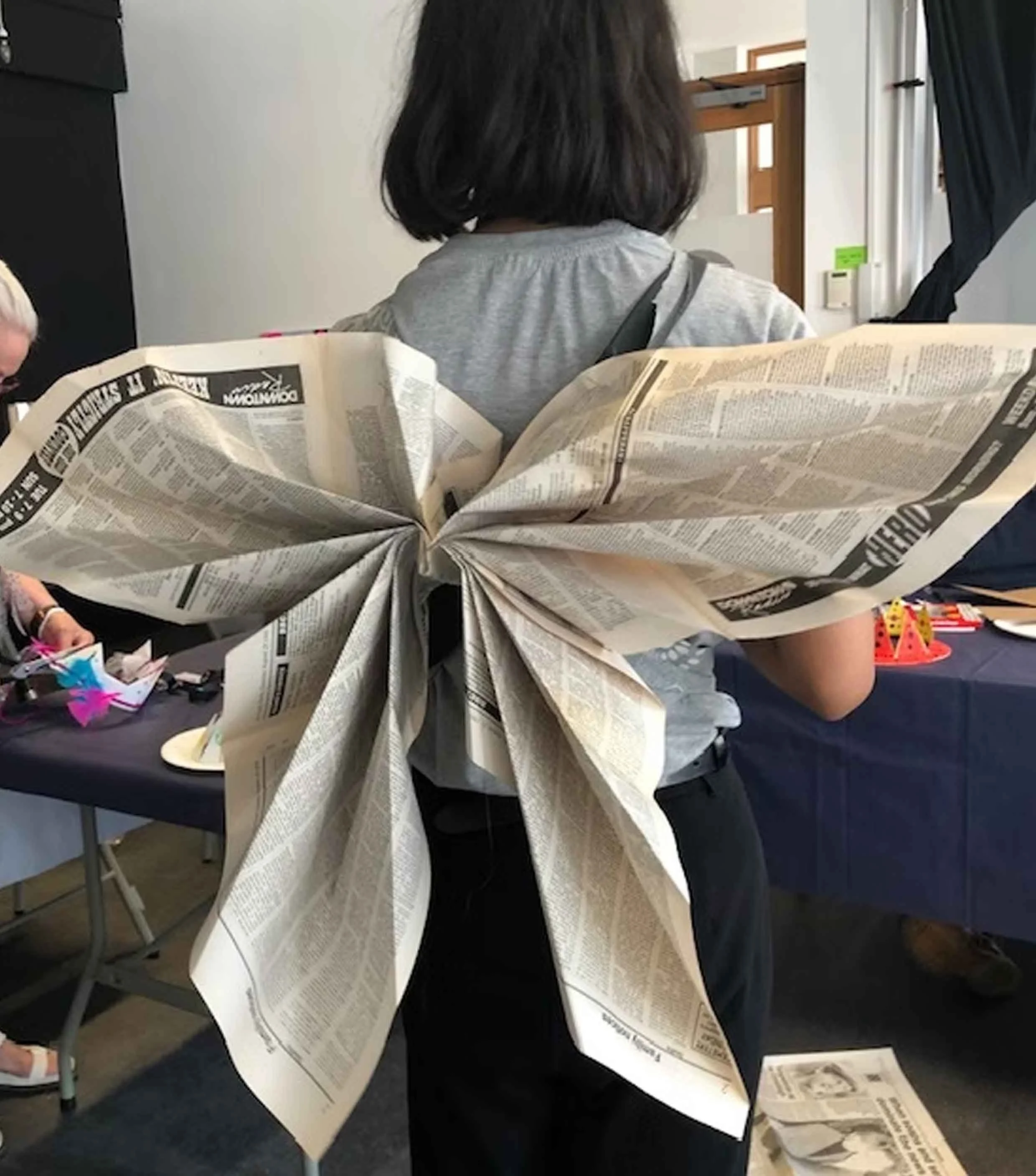 Person standing with newspaper garment in a room with tables and chairs, other people, and a doorway in the background.