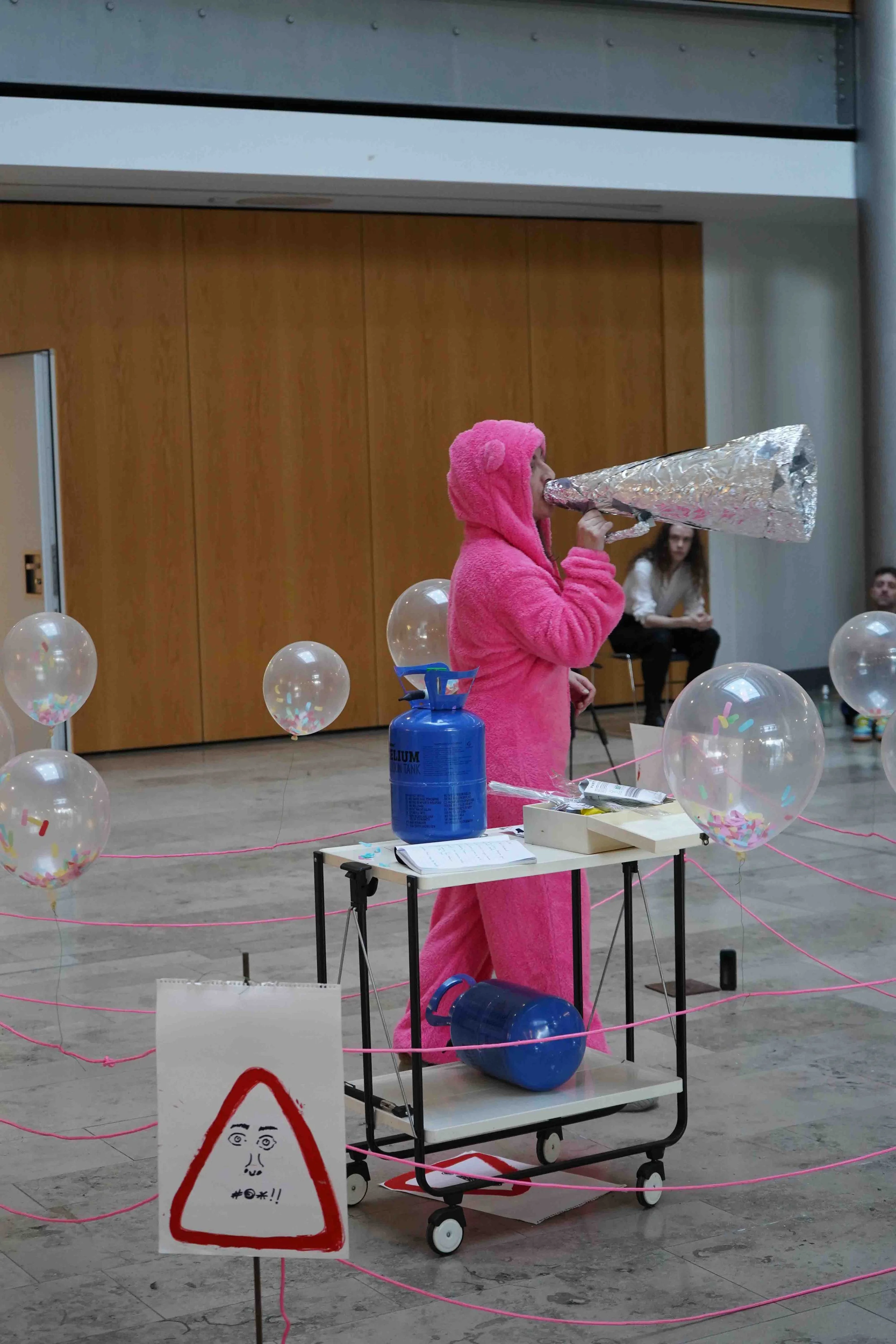 A person in a pink bear onesie costume shouting through a tinfoil megaphone. The scene is decorated with balloons and pink ribbons, and there are people sitting in the background.