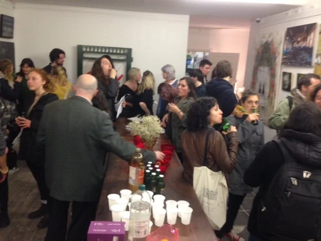 People attending an art gallery event, socializing and viewing artwork on the walls, with drinks and snacks on a table.