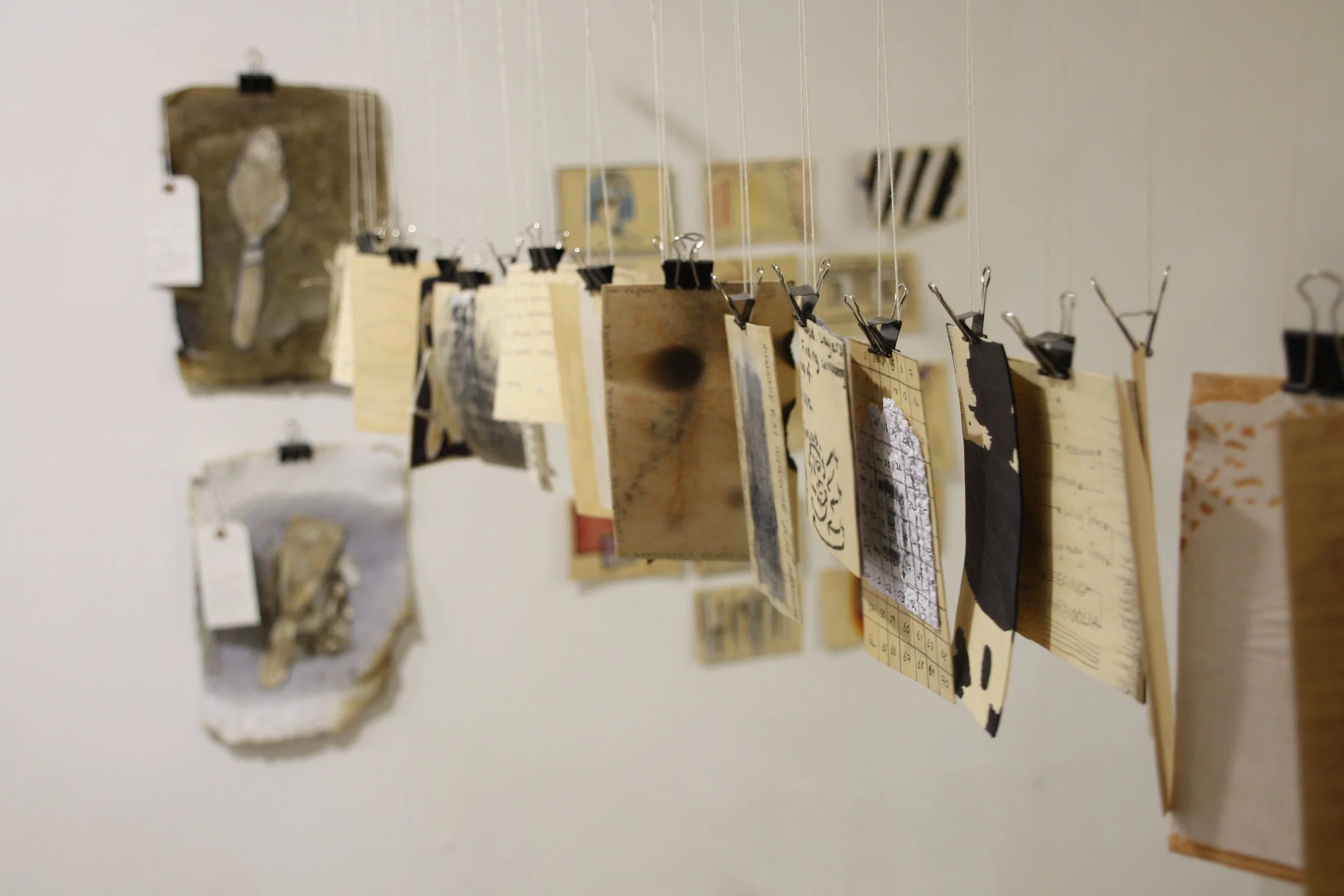 A series of small paper notes, photographs, and cards are clipped to thin strings with black binder clips, hanging against a light-colored wall. The notes appear to be handwritten or printed and are unevenly spaced.