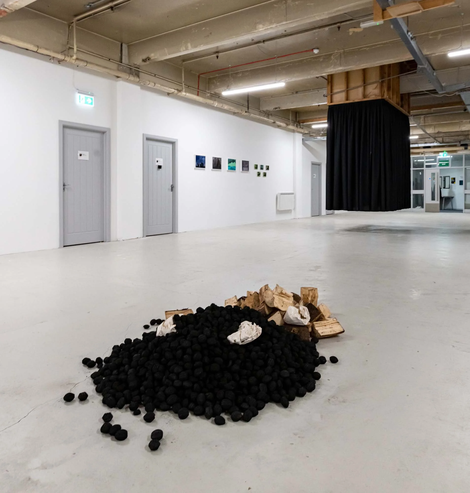 Interior of an art gallery with minimalistic white walls, doors, ceiling with exposed piping, and a fire exit sign. A black curtain hangs from the ceiling, and a small pile of wood and stones is on the floor in front of the camera. Wall art is displayed on the white wall.