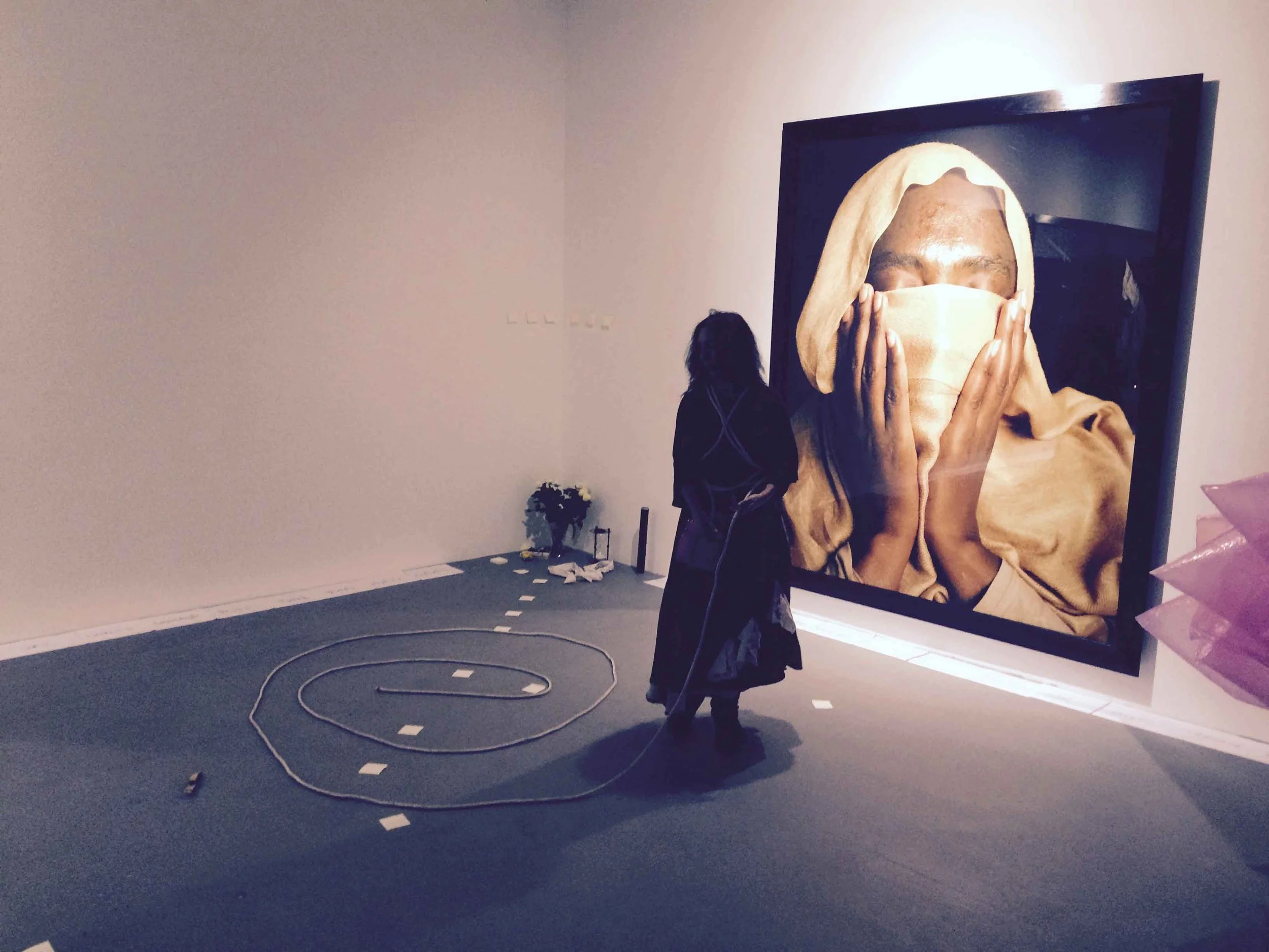 An art exhibition features a large portrait of a person covering their face with their hands. The room has minimal decor, with a small table and some objects on the far side. The floor has a circular track with road markings, possibly for an interact