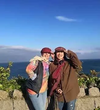 Two women standing outdoors near the ocean on a sunny day, both wearing sunglasses and hats, smiling at the camera.