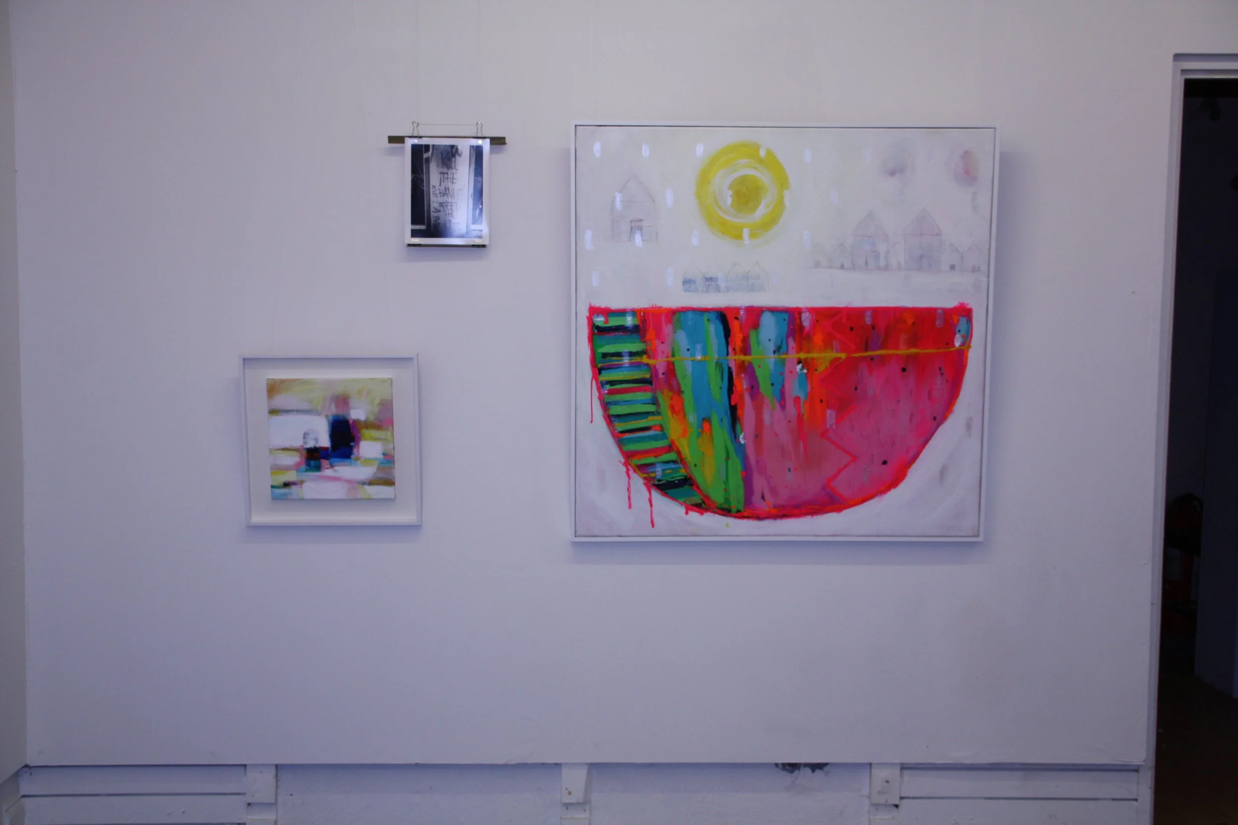 Gallery wall with abstract art pieces, including a colorful geometric painting, a sketch of houses under a yellow sun, and a small digital photograph.