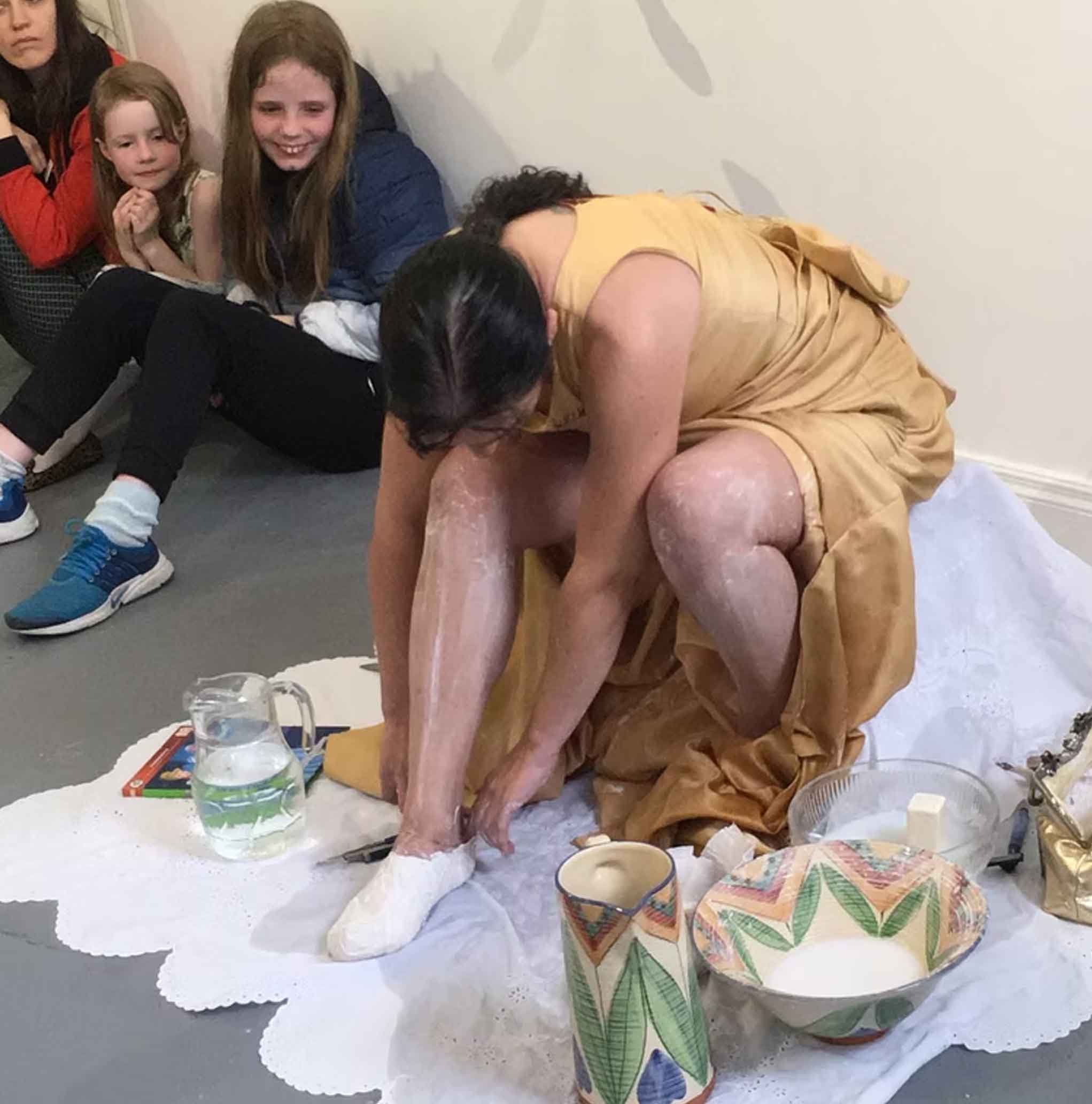 Group of people around a clay or ceramic sculpture of a kneeling woman in a yellow dress, with a large green leaf pattern on her skirt, and a head covering, working in an art studio or classroom with pottery and art supplies.