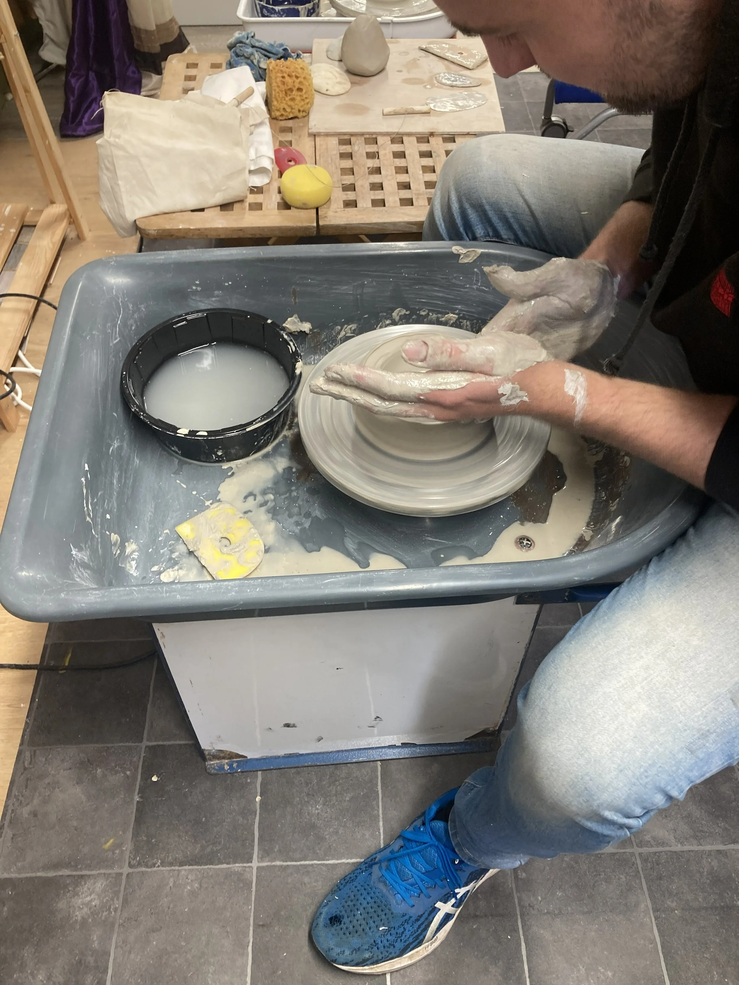person sitting at a pottery wheel throwing a small pot with clay covered hands, light blue jeans and blue trainers