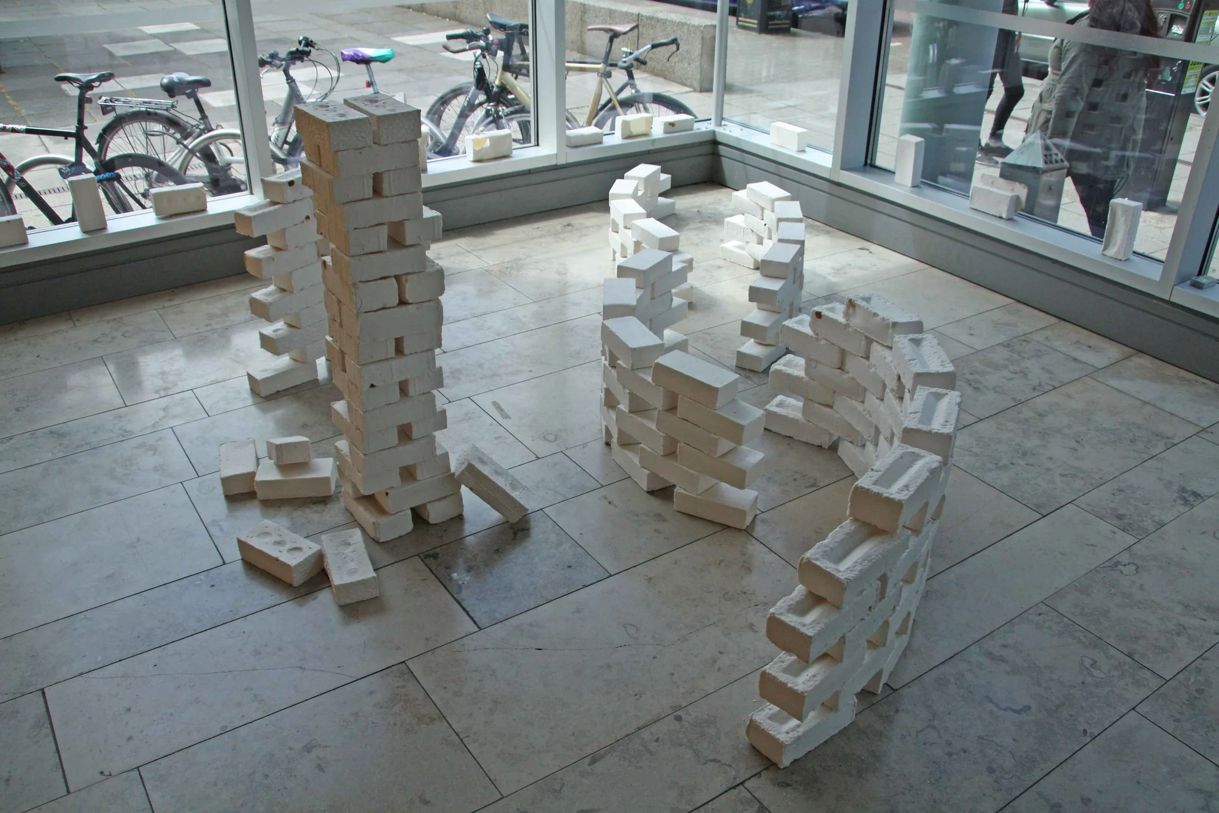 Indoor display of white bricks arranged in tall stacks and several structures, with large glass windows showing bicycles parked outside and people walking by.