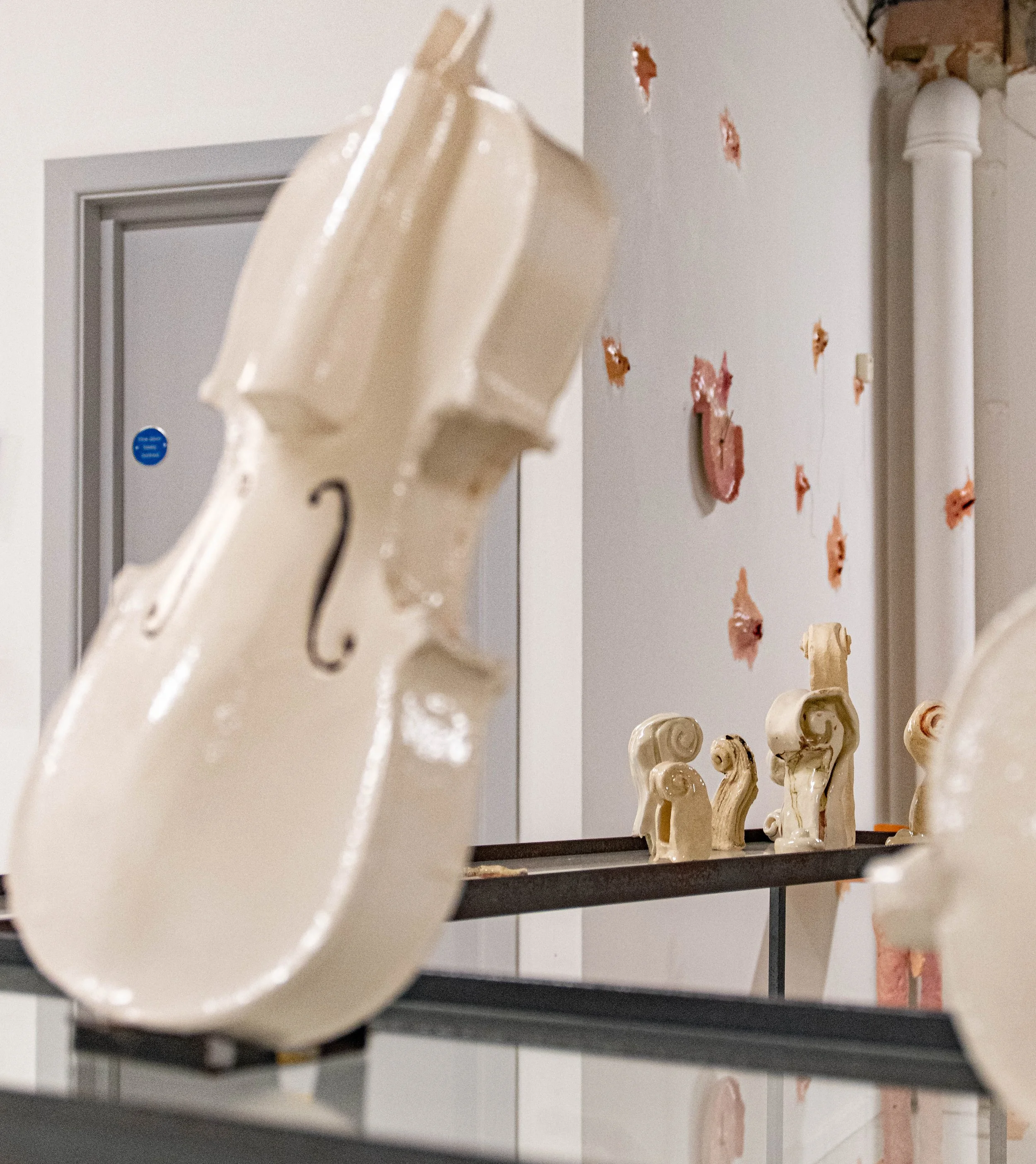 Close-up of a white ceramic violin in focus, with a display of ceramic art pieces on a black shelf and a decorated wall with orange and red paper flowers in the background.