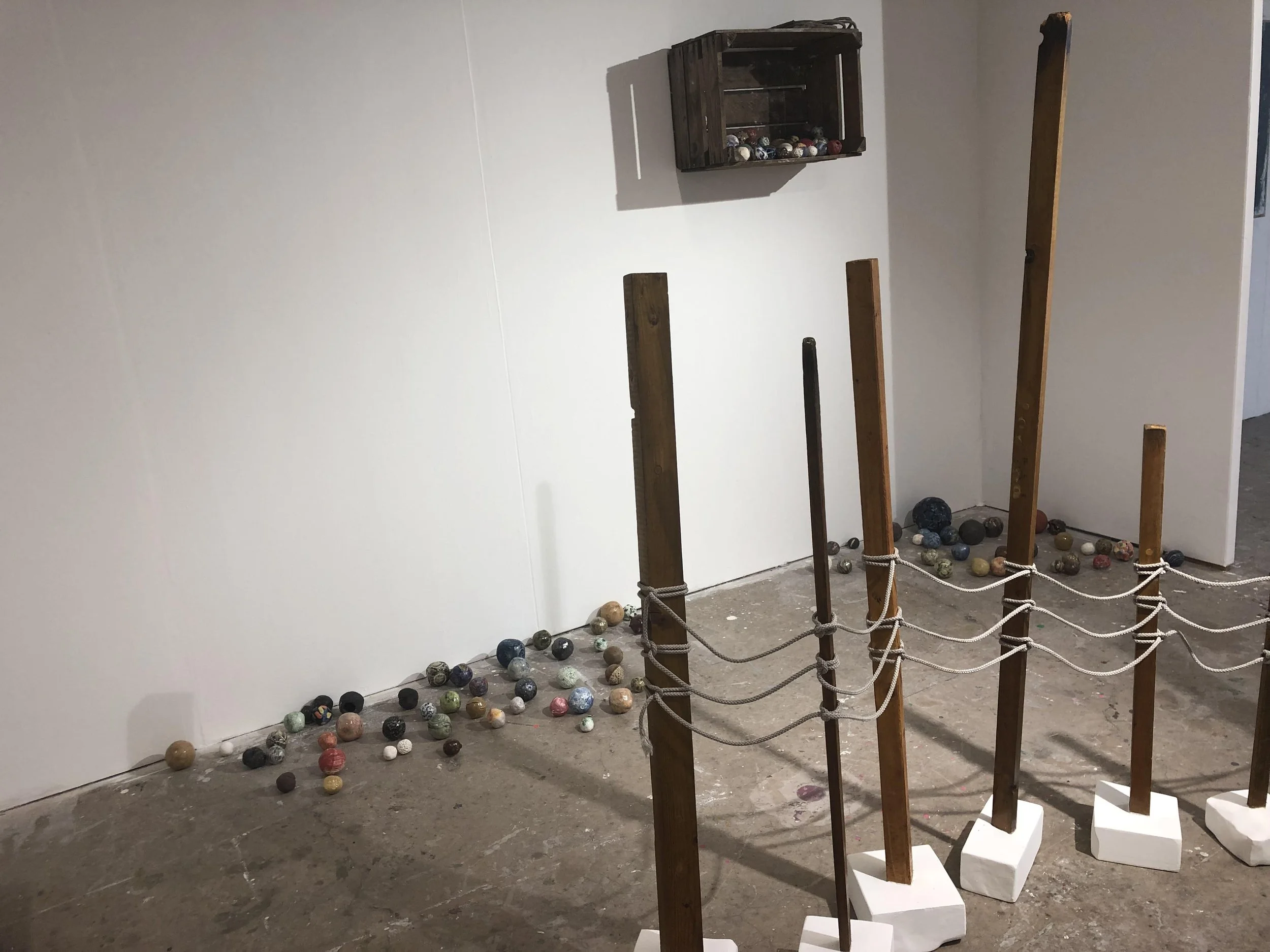 Art installation with wooden posts, white bases, and numerous marbles on the floor, with a small display case on the white wall in a gallery setting.