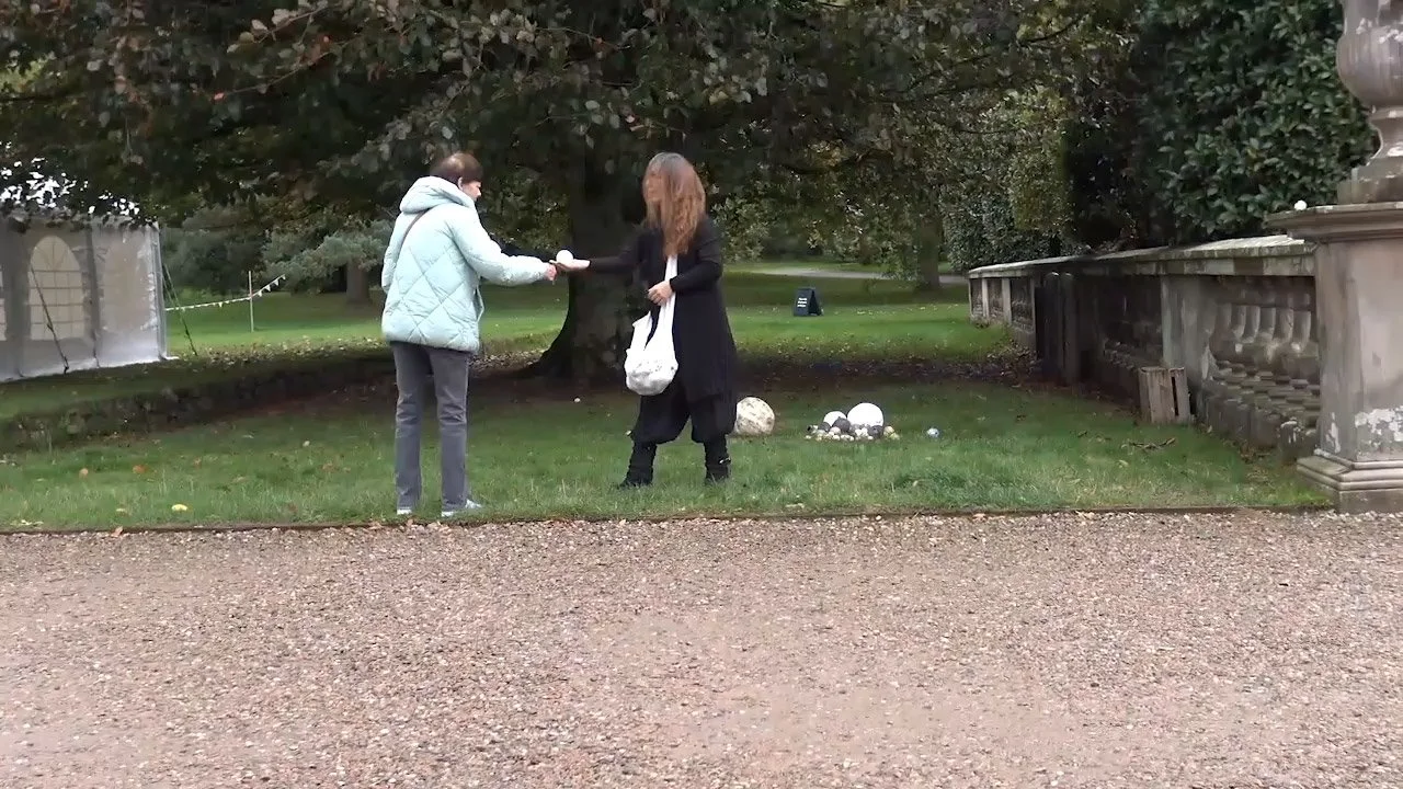 A person in a gray jacket handing something to a woman with long hair, dressed in black, outside in a park with grass and trees.