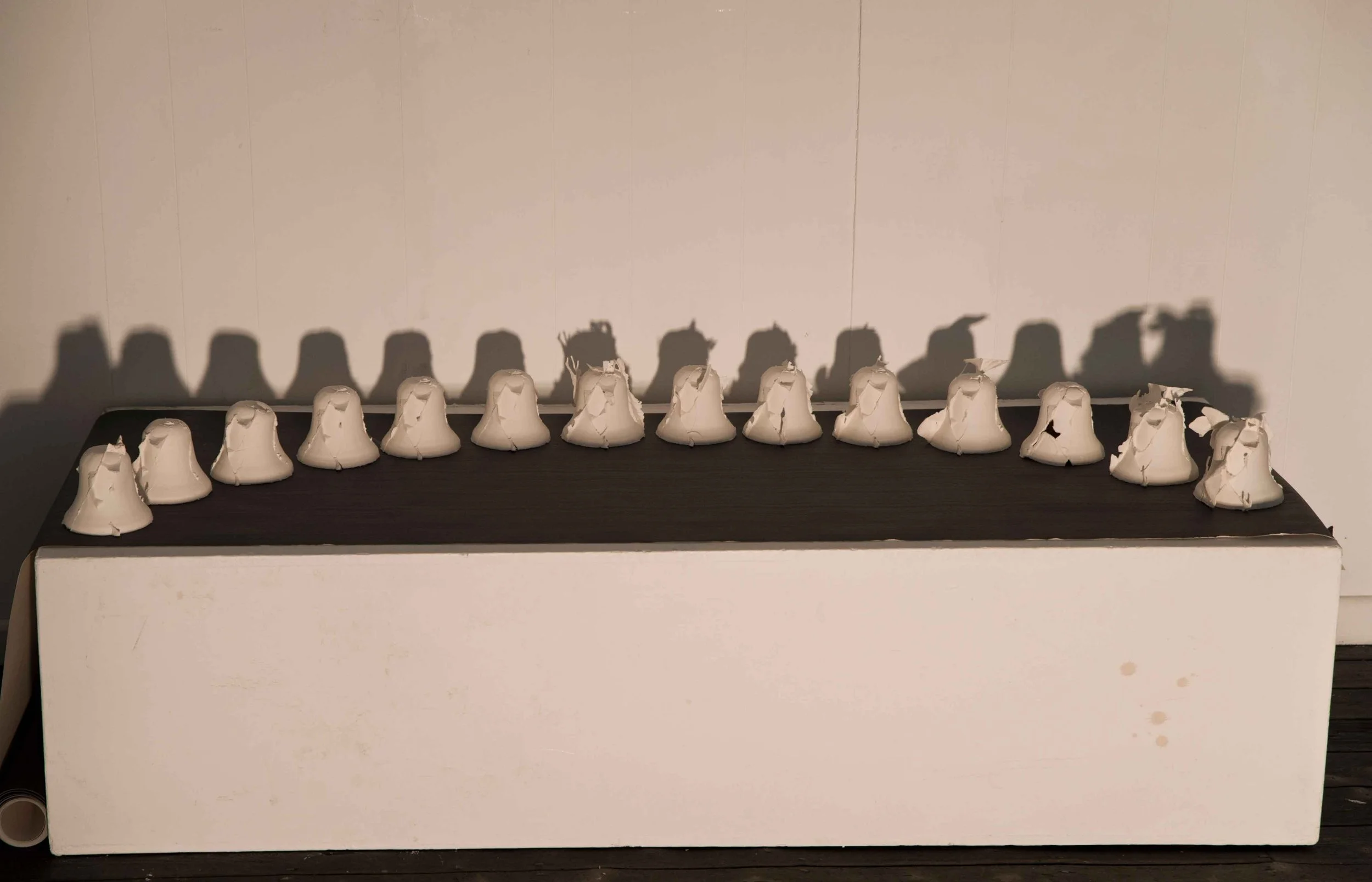 A line of crumpled white bells arranged on a dark surface with a white background, casting shadows on the wall behind.