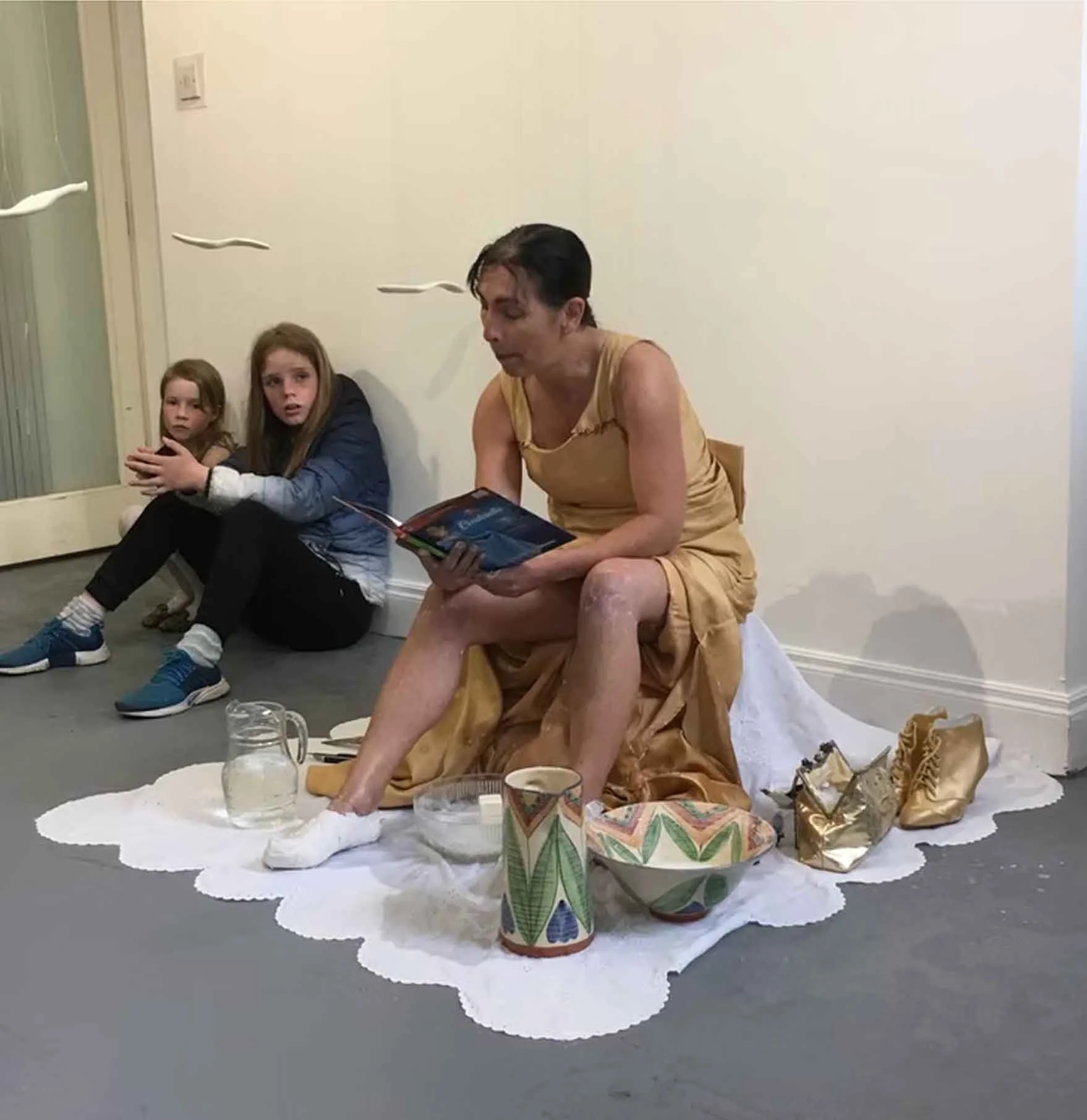 A woman sitting on a white sheet with her feet on the floor, reading a magazine, and two young girls sitting beside her with one of the girls hugging the other. There are various objects on the sheet, including paper cups, a glass jar, and a wrapped 