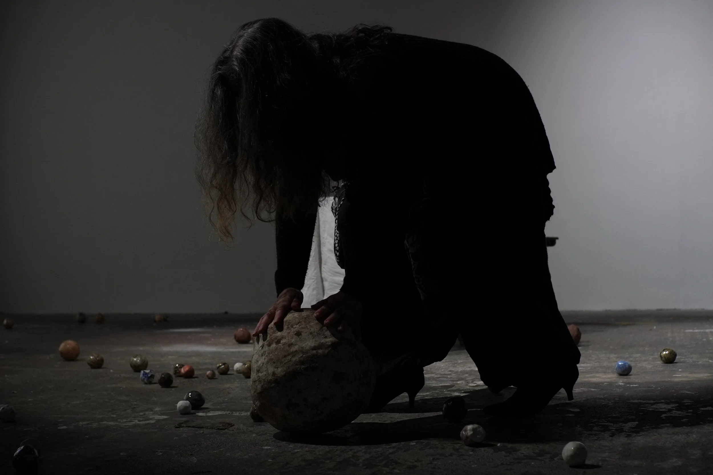 A person with long hair, dressed in dark clothing, kneeling and touching a large rock surrounded by scattered colorful balls in a dimly lit room.