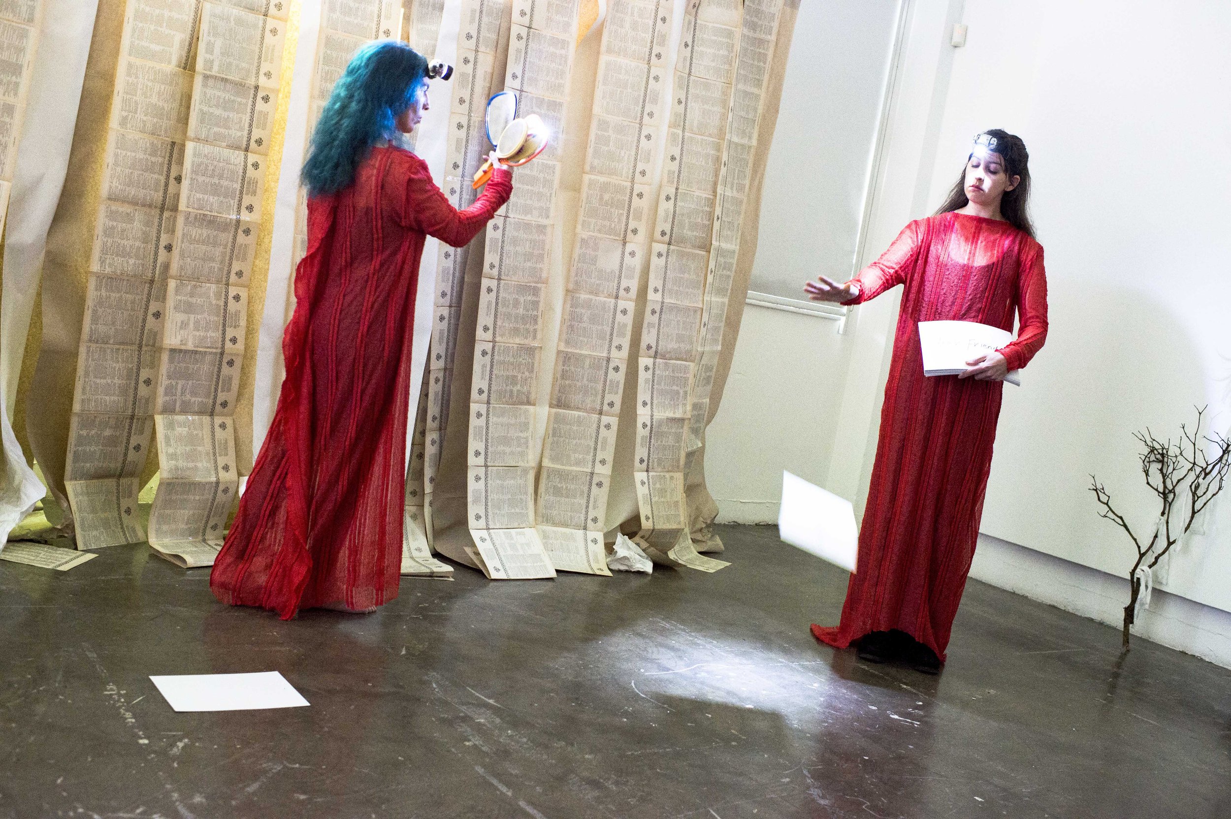 Two women in red dresses are performing in an art installation with large pages of text hanging from the ceiling. One woman is holding a book and the other appears to be reading or presenting, both wearing headlamps.