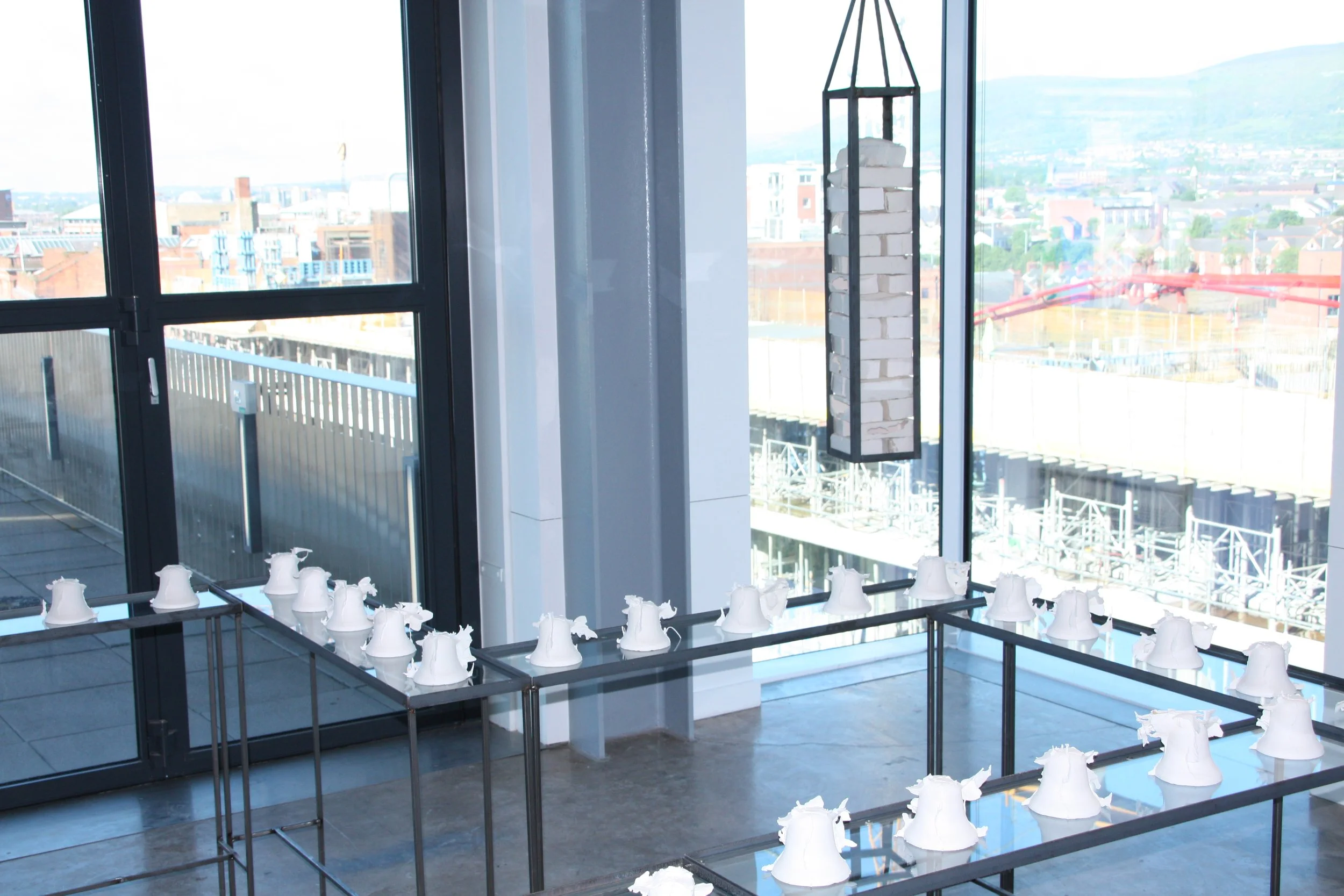 Interior of a building with large floor-to-ceiling windows showing a cityscape and mountains in the distance. There are multiple white ceramic bell sculptures arranged on black metal stands in the foreground.