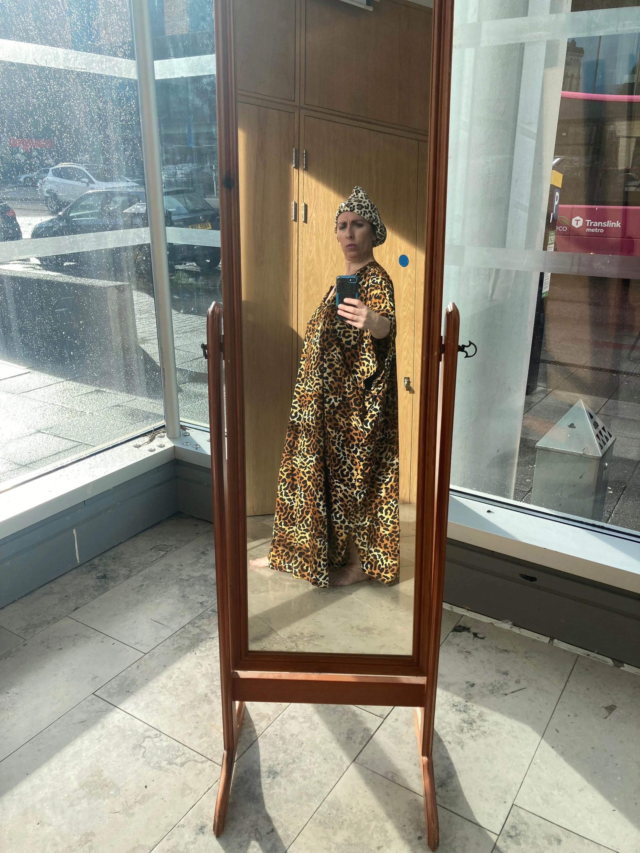 Woman dressed in leopard print clothing standing in front of a full-length mirror, taking a selfie in glass walled gallery space, with a wooden cabinet and a rainy street scene visible outside the window.