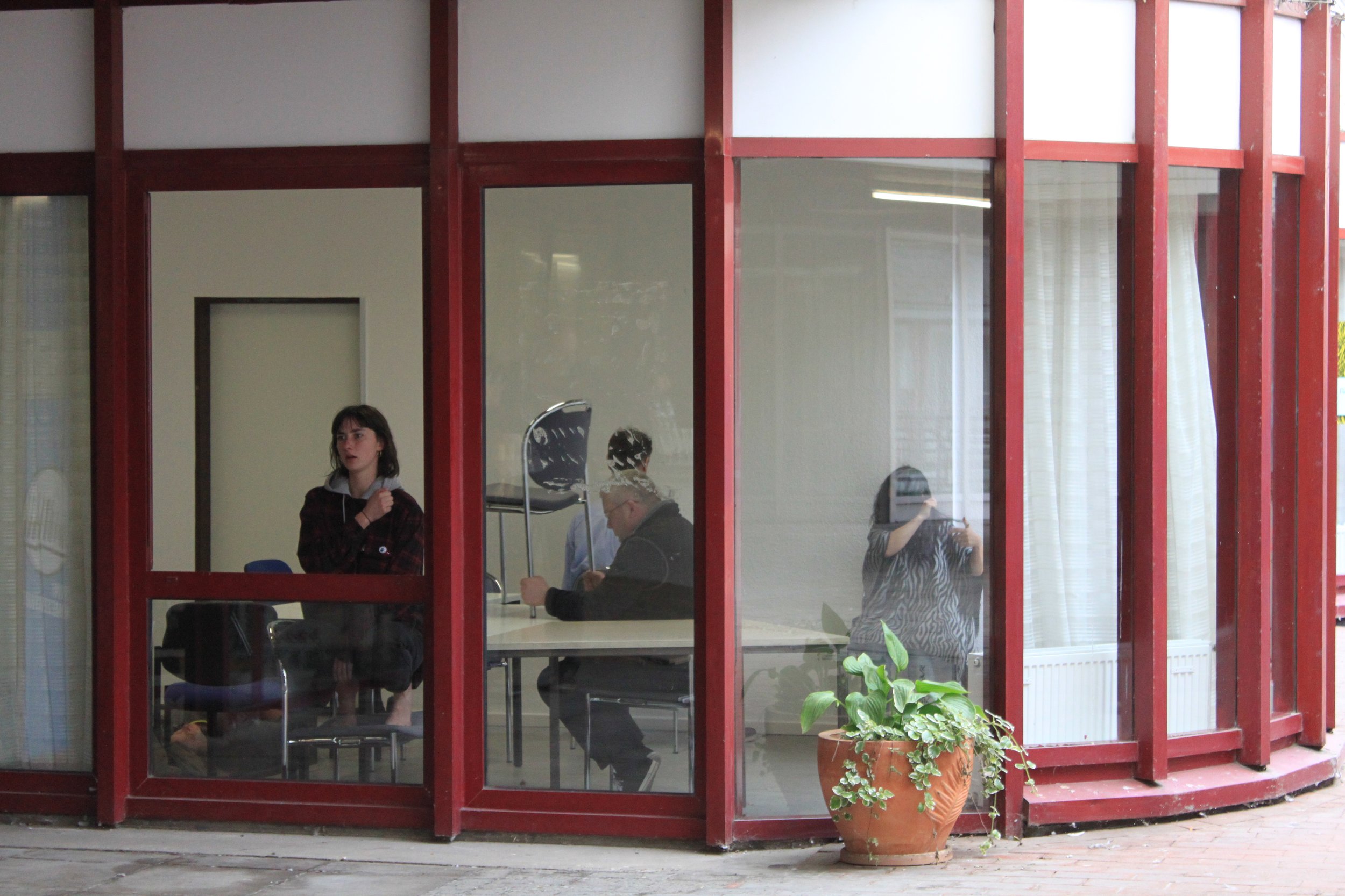 People sitting and talking inside a room viewed through large glass windows with a red frame, outside a potted plant is visible.
