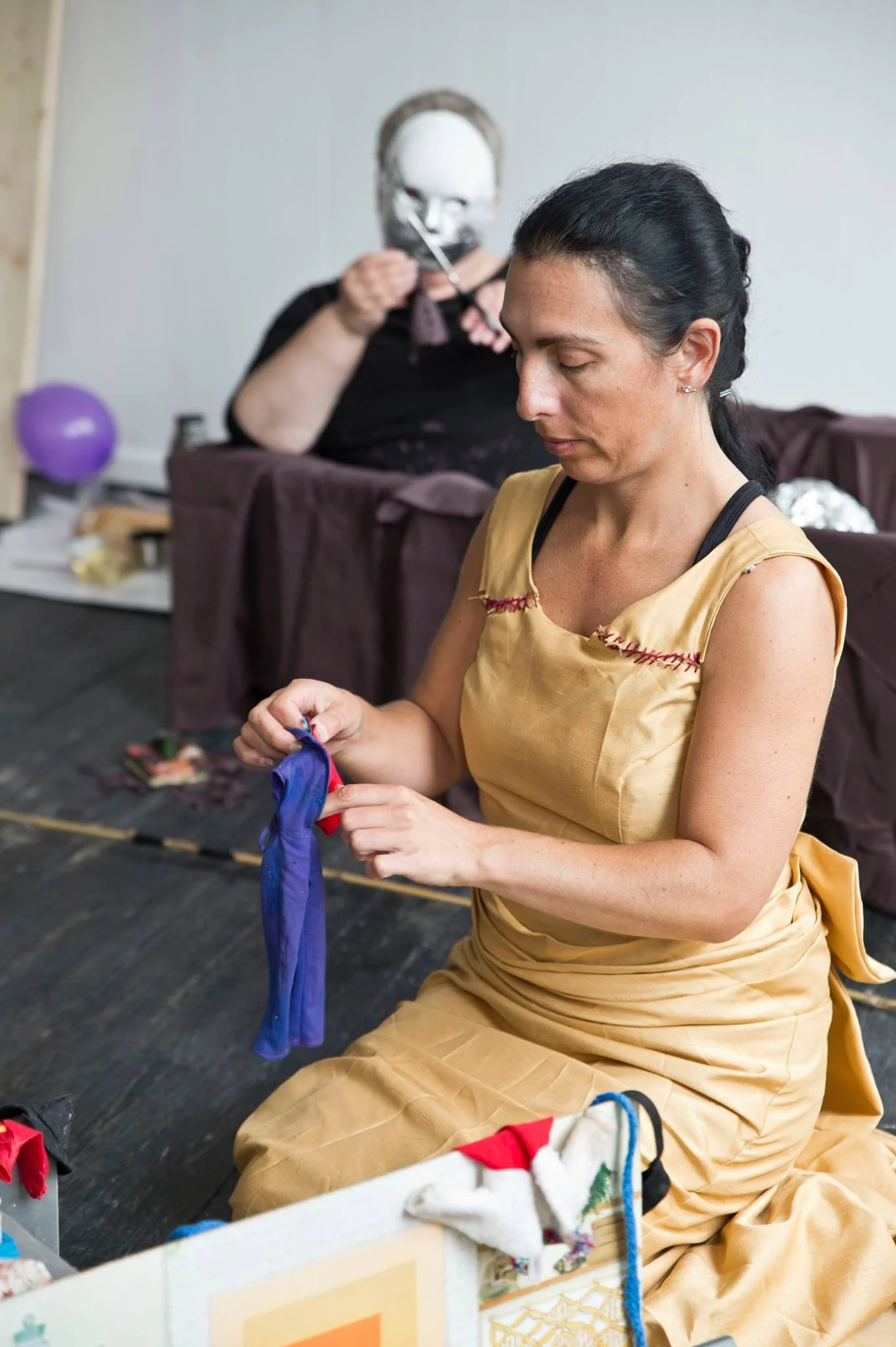A woman in a yellow dress sitting on the floor, sewing a piece of fabric, with a woman in the background wearing a mask and holding a tool near her face.
