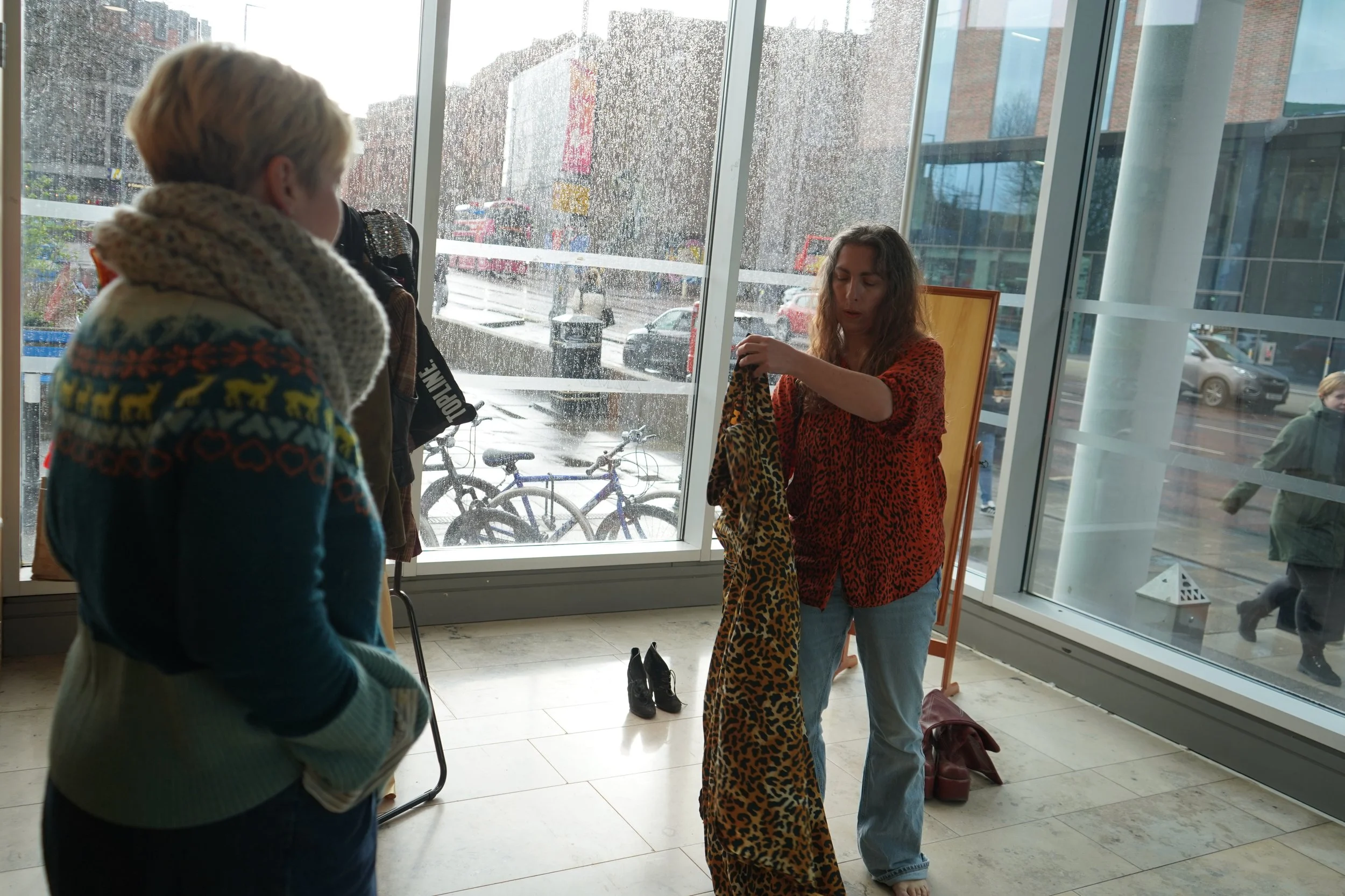 Three women inside a modern building near large glass windows, looking at and discussing animal-print clothing. The outside scene shows bicycles parked and rainy weather.