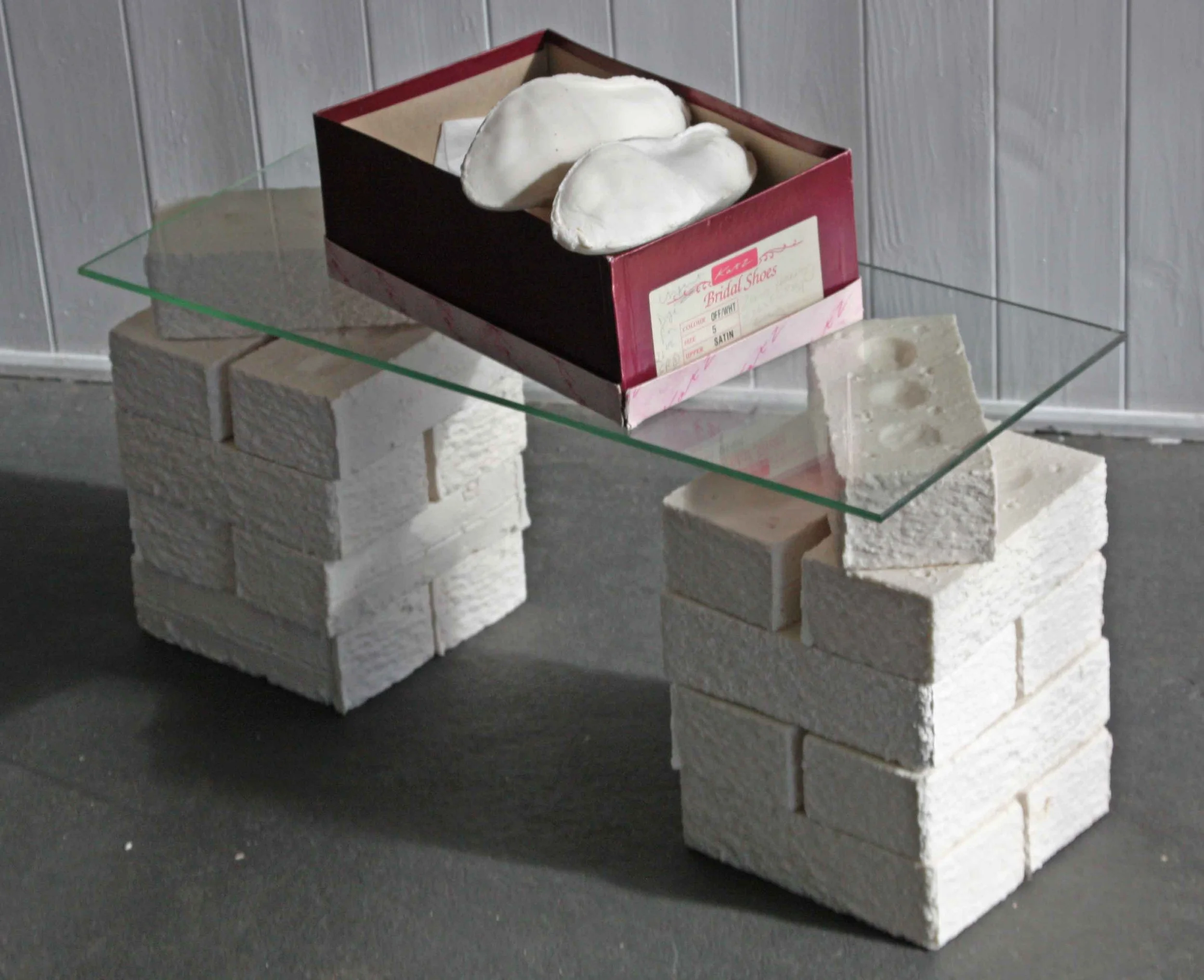 A small box labeled "Bridal Shoes" contains two white shoes and is placed on a glass shelf supported by white bricks, against a background of wooden wall panels.