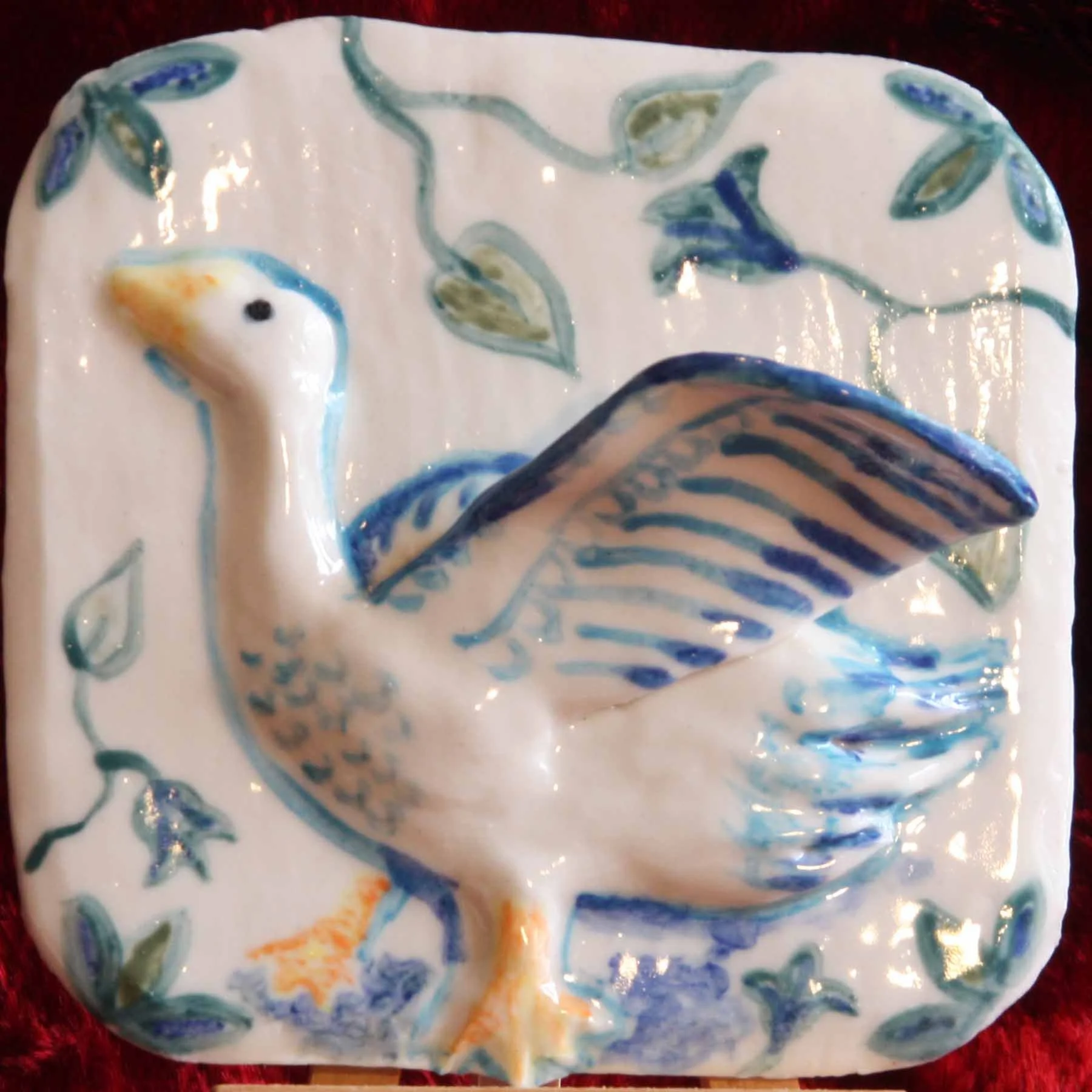 A decorative ceramic plate with a goose and blue and green leaf and vine patterns, featuring a three-dimensional fish sculpture protruding from the surface, set against a red background.