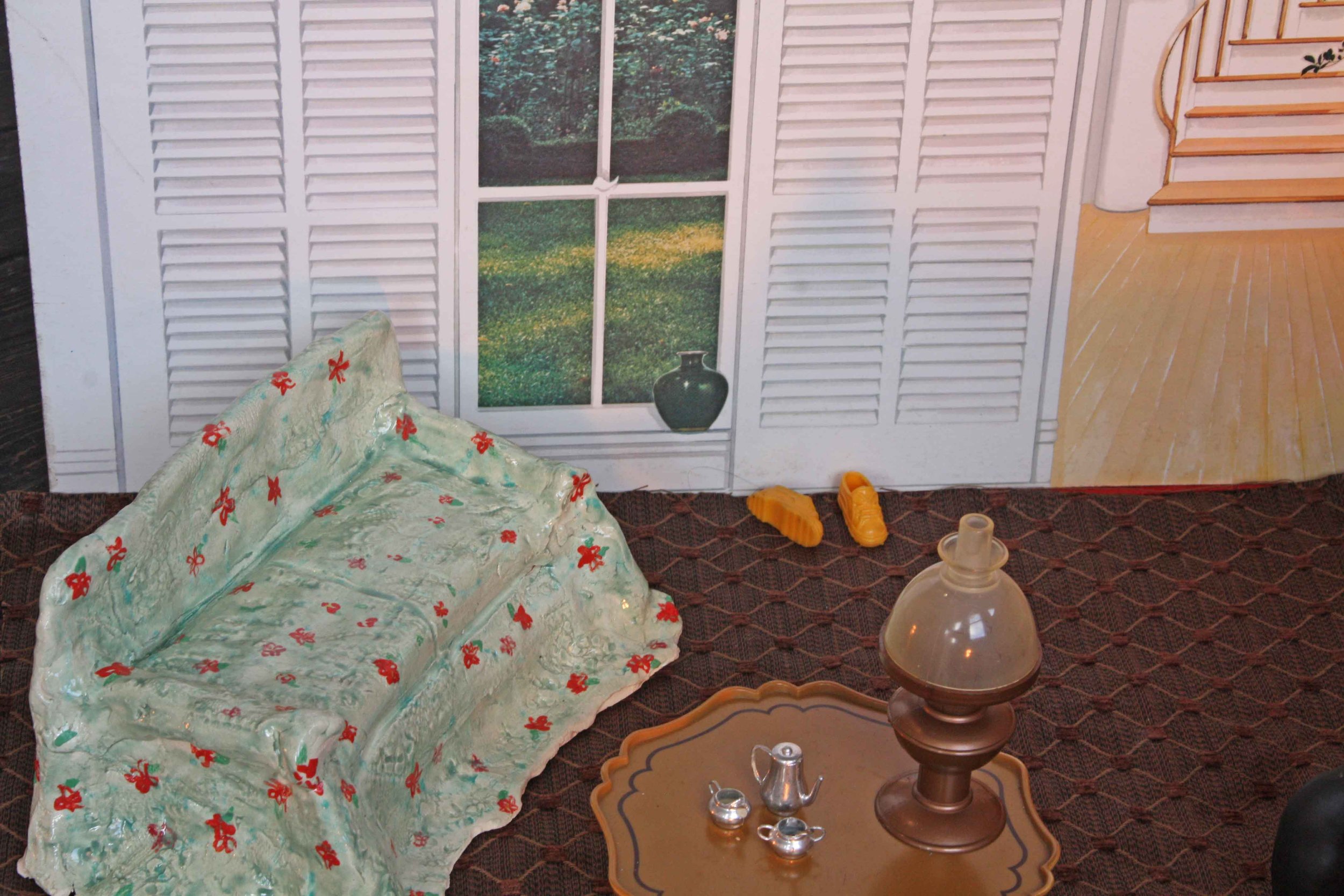 A miniature room scene with a window, white shutters, and a view of greenery outside. There is a floral-patterned cloth on the left, a small copper-colored table with a glass top, and various small decorative items on the table including a small teap