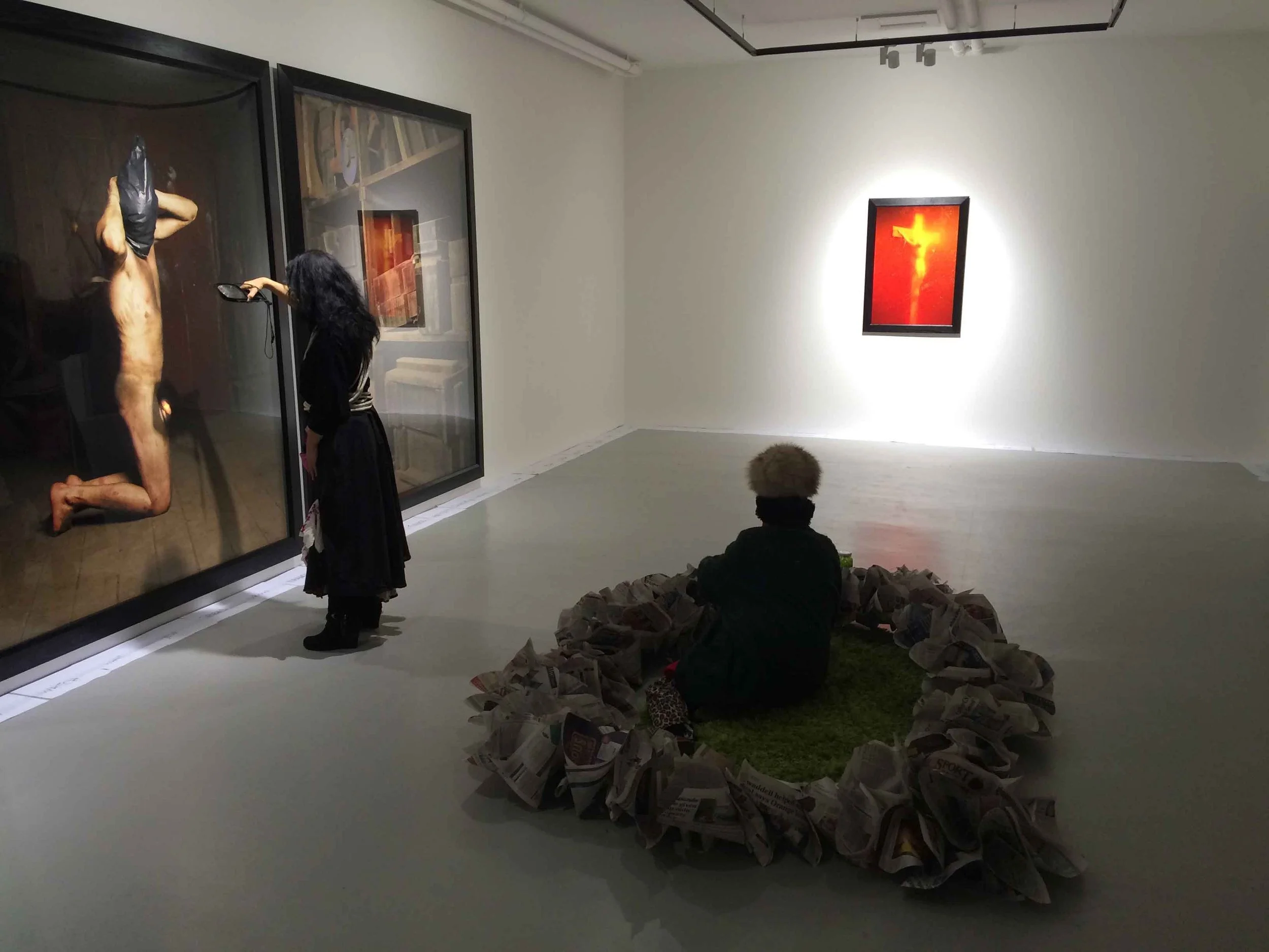 An art gallery with a large painting of a crucifixion scene on the wall, and a woman viewing it closely. There are two people seated on the floor surrounded by a large pile of newspapers, and a smaller framed artwork with a neon cross on the wall.
