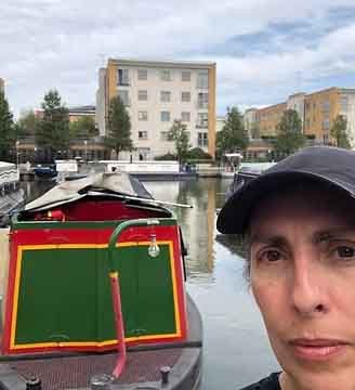 A person wearing a cap takes a selfie near a marina with boats and a building in the background.