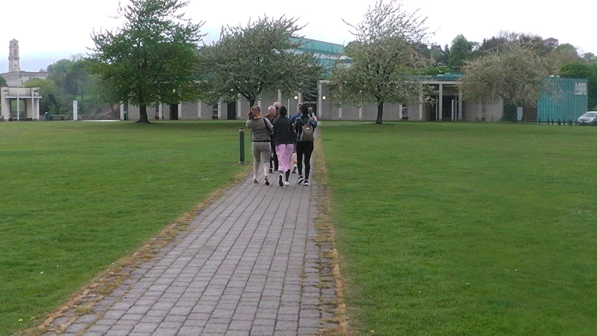 A group of six people walking down a paved path in a grassy park with trees and a building in the background.
