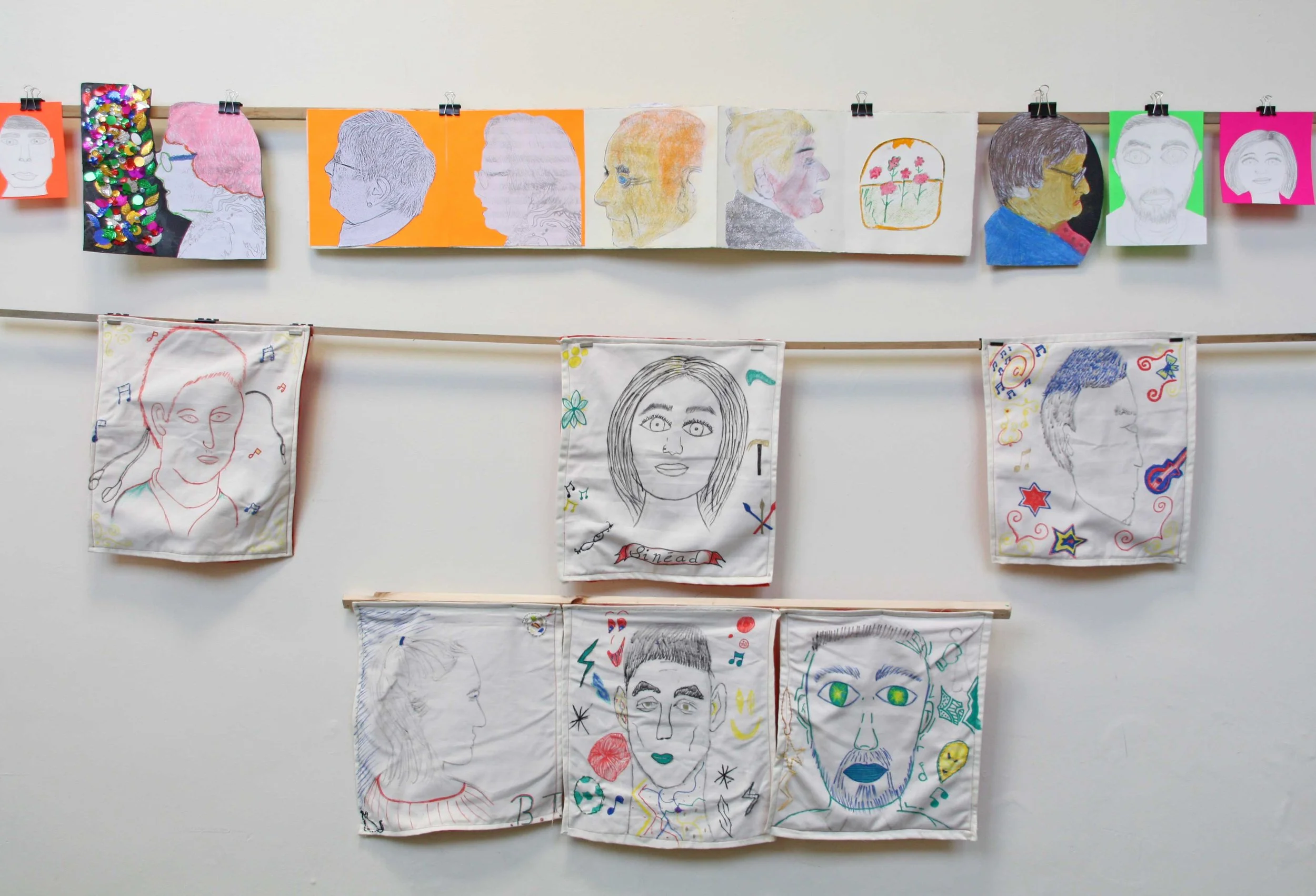 Displays a collection of portrait drawings and illustrations arranged on two horizontal hooks, featuring various artistic styles and colorful details, including sketches, painted portraits, and decorative fabric art.