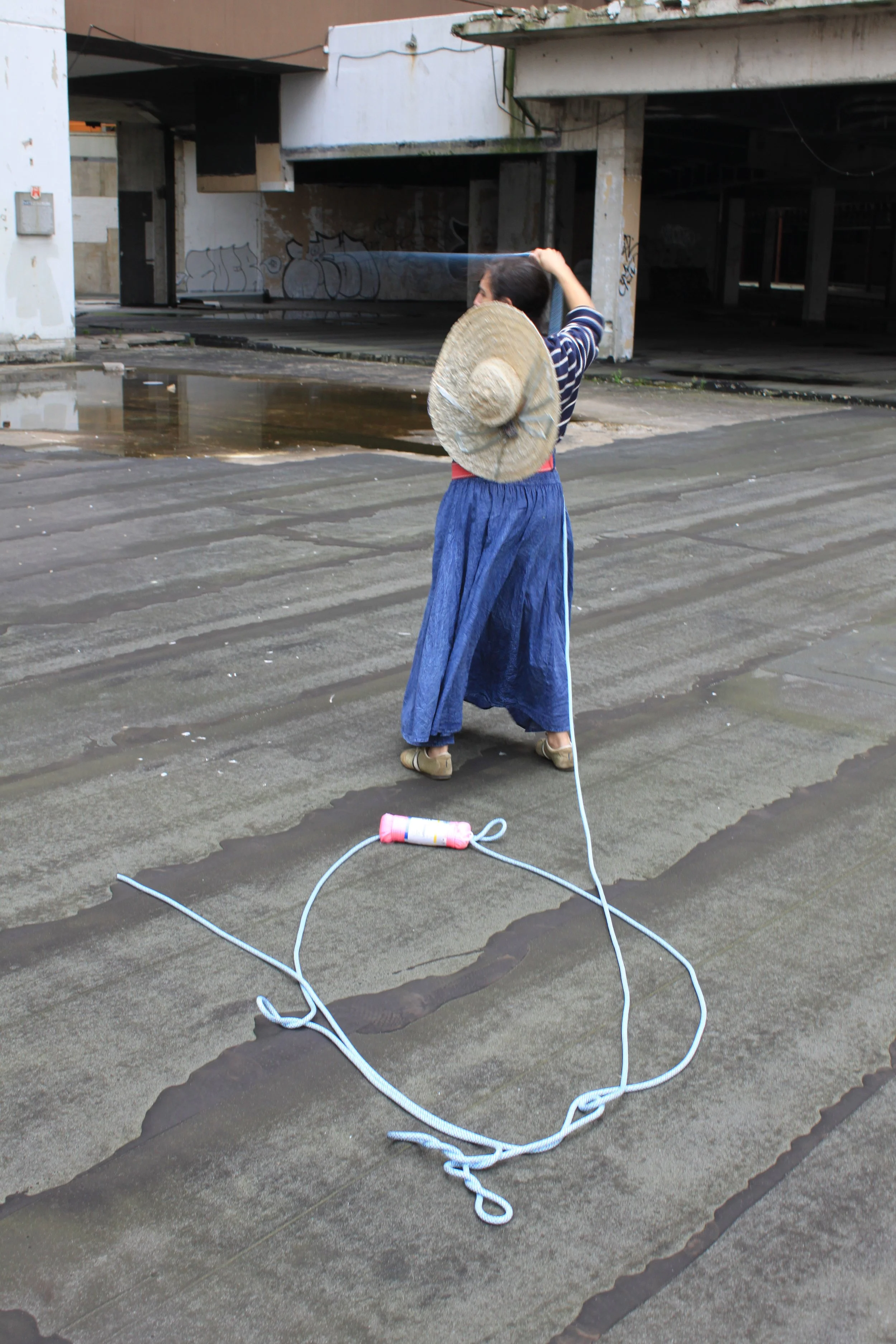 Person wearing a straw hat and a striped shirt standing in an urban parking lot, holding a sprayer, with a long hose on the ground and a large puddle of water nearby.