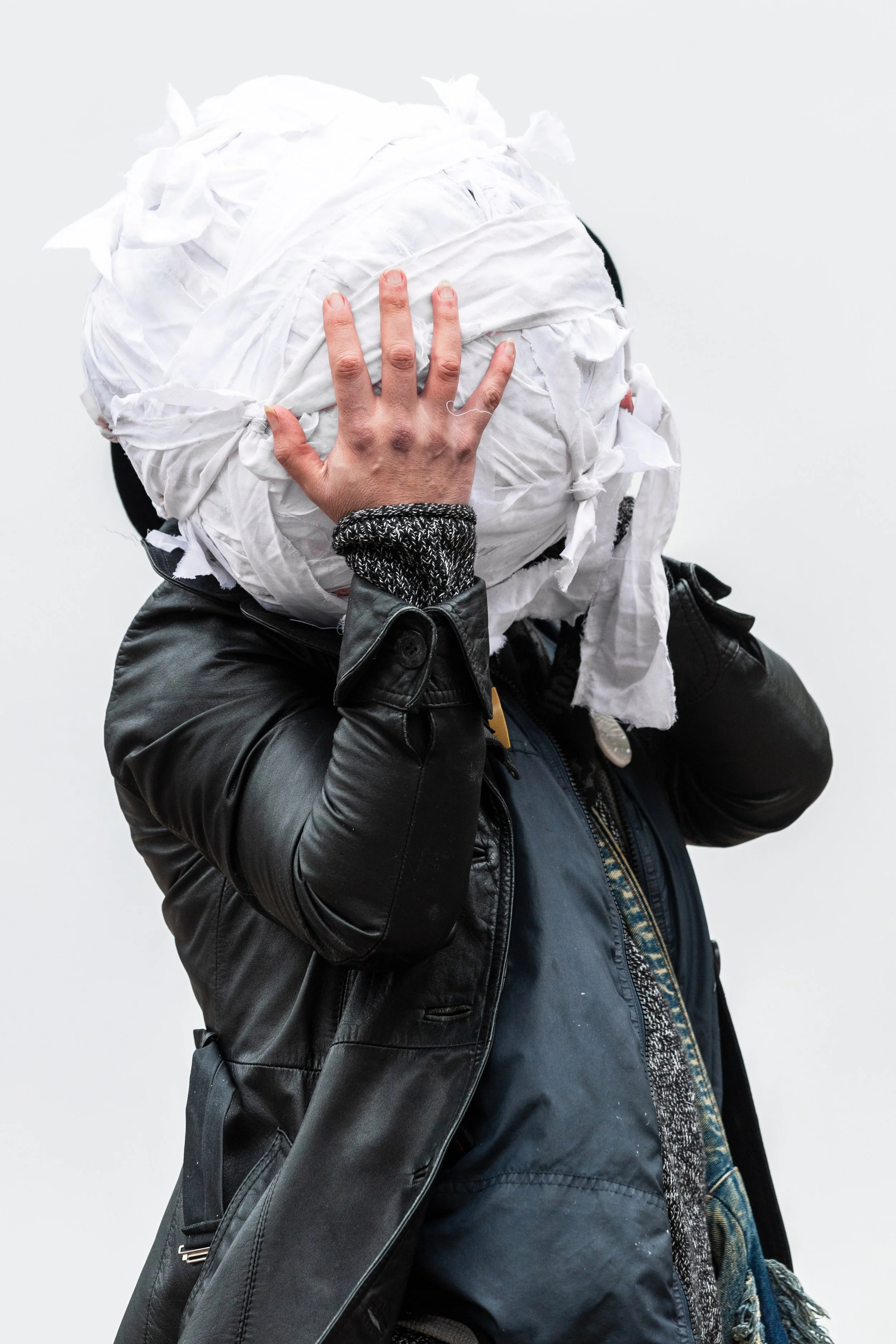 Person wearing a black leather jacket holding their head with a large bundle of white cloth or paper covering their face.