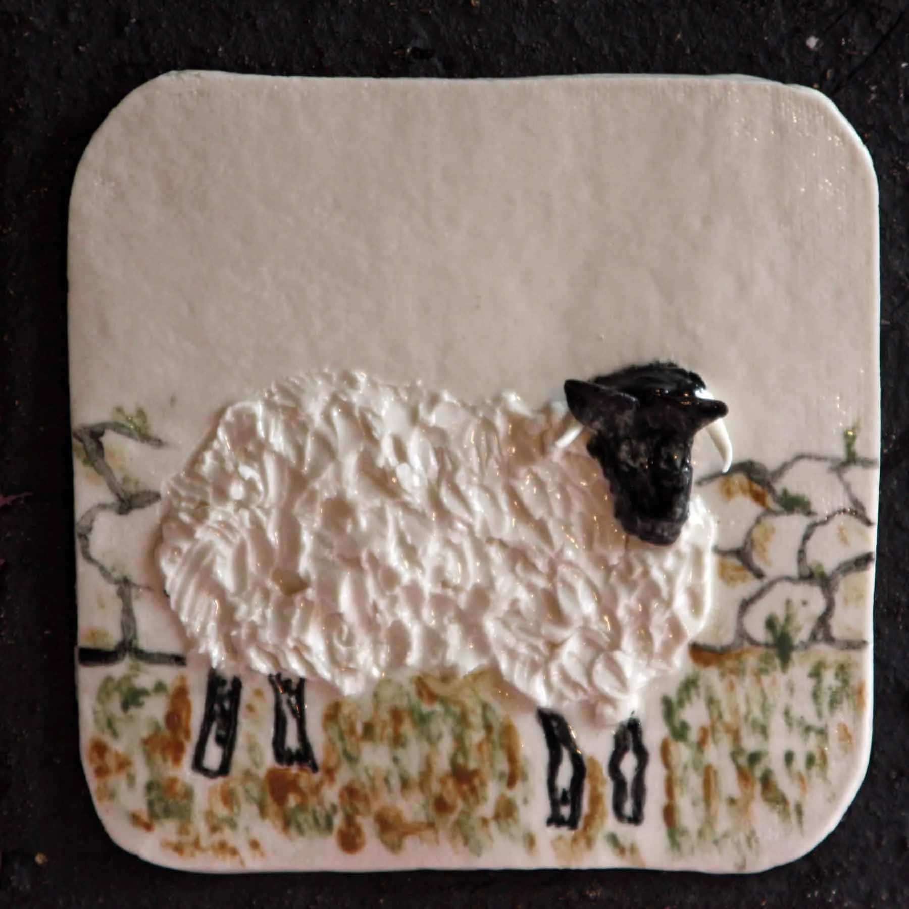 Decorative rectangular tile with a white background, painted floral designs in green, orange, and brown, topped with a sheep.