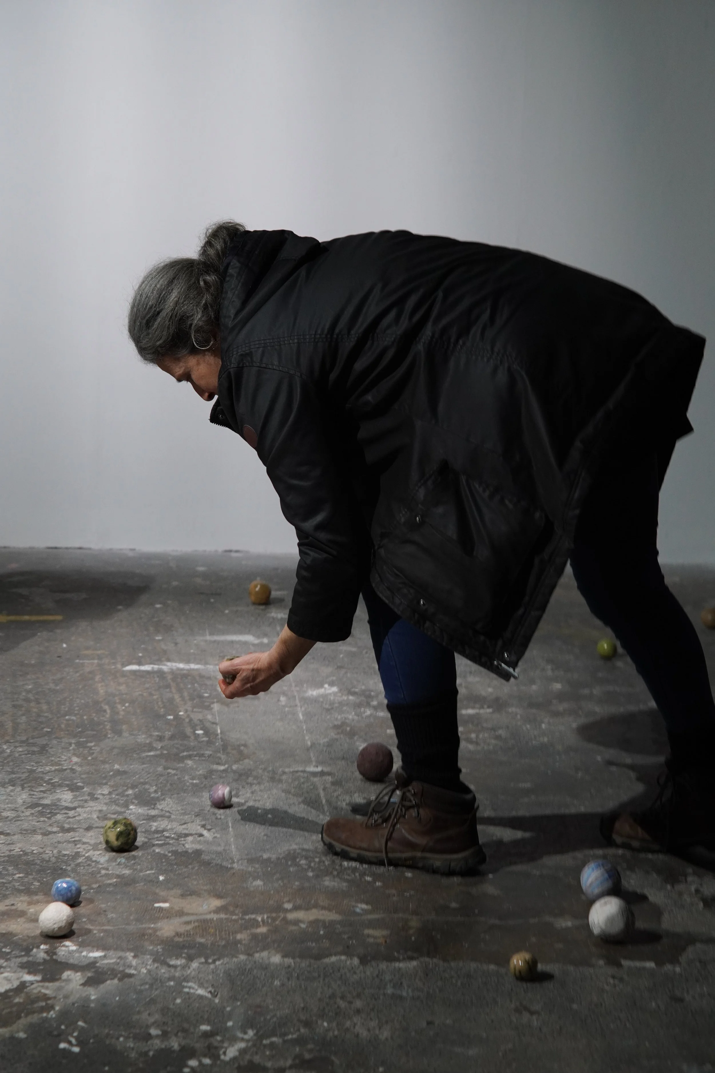 A person bending over to pick up a colorful marble from a textured, dark floor, with several other marbles scattered around. The person is wearing a black jacket, blue jeans, and brown boots, and has gray hair. The background features a plain, light-