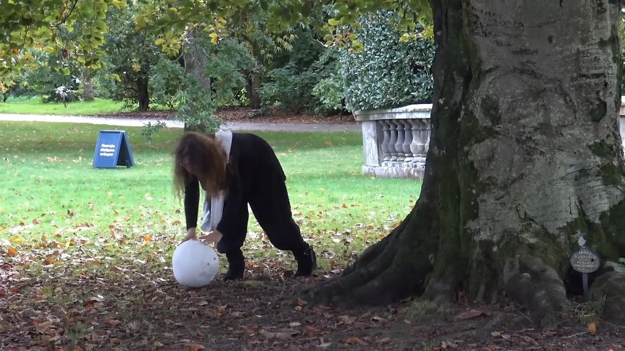 A woman in a black coat and white shirt is outdoors, bending over to pick up or interact with a white sphere on the ground, near the base of a large tree with textured bark.
