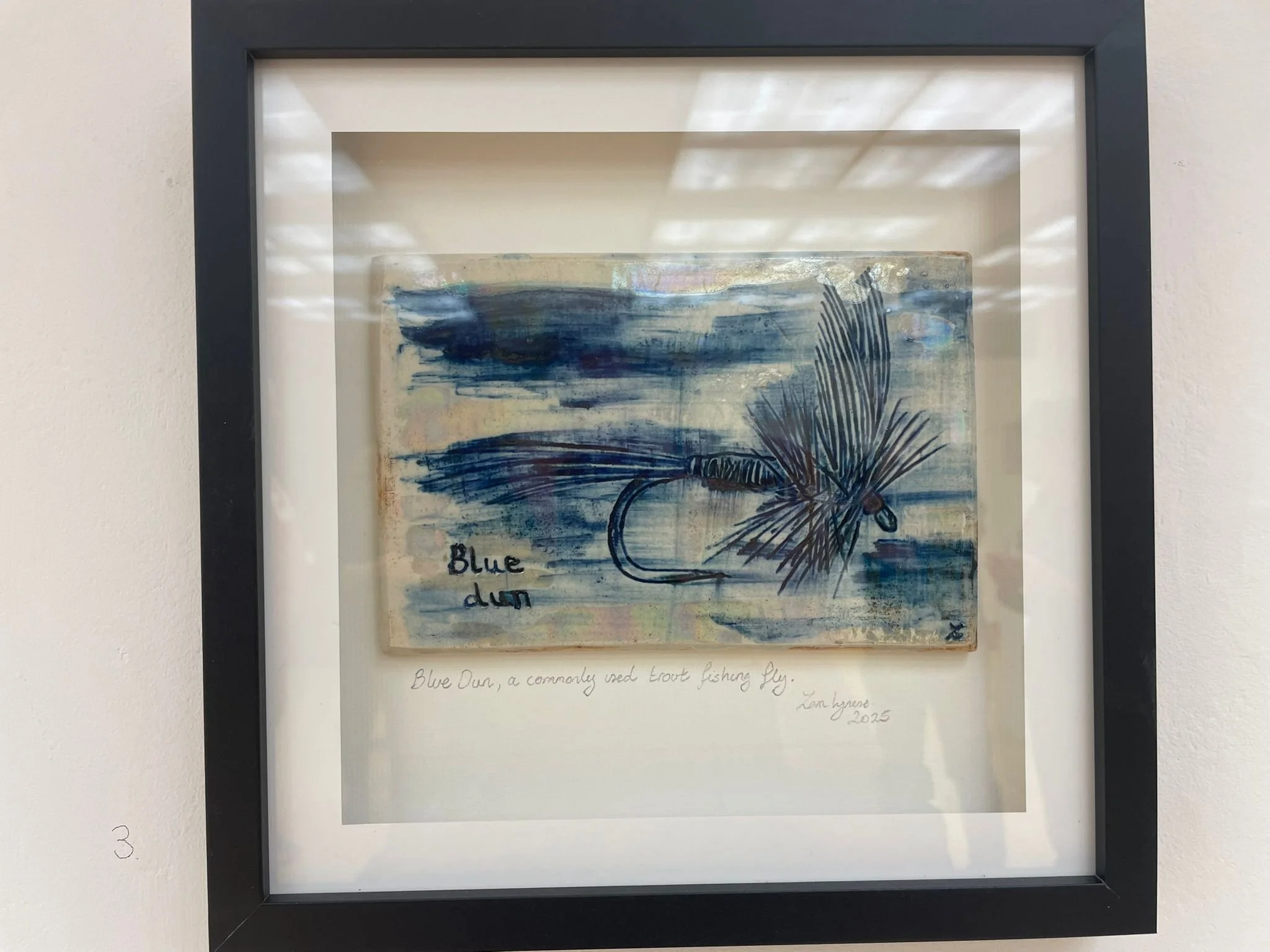 Framed ceramic fishing fly with a blue ocean wave and hand written text 'Blue' on the artwork.