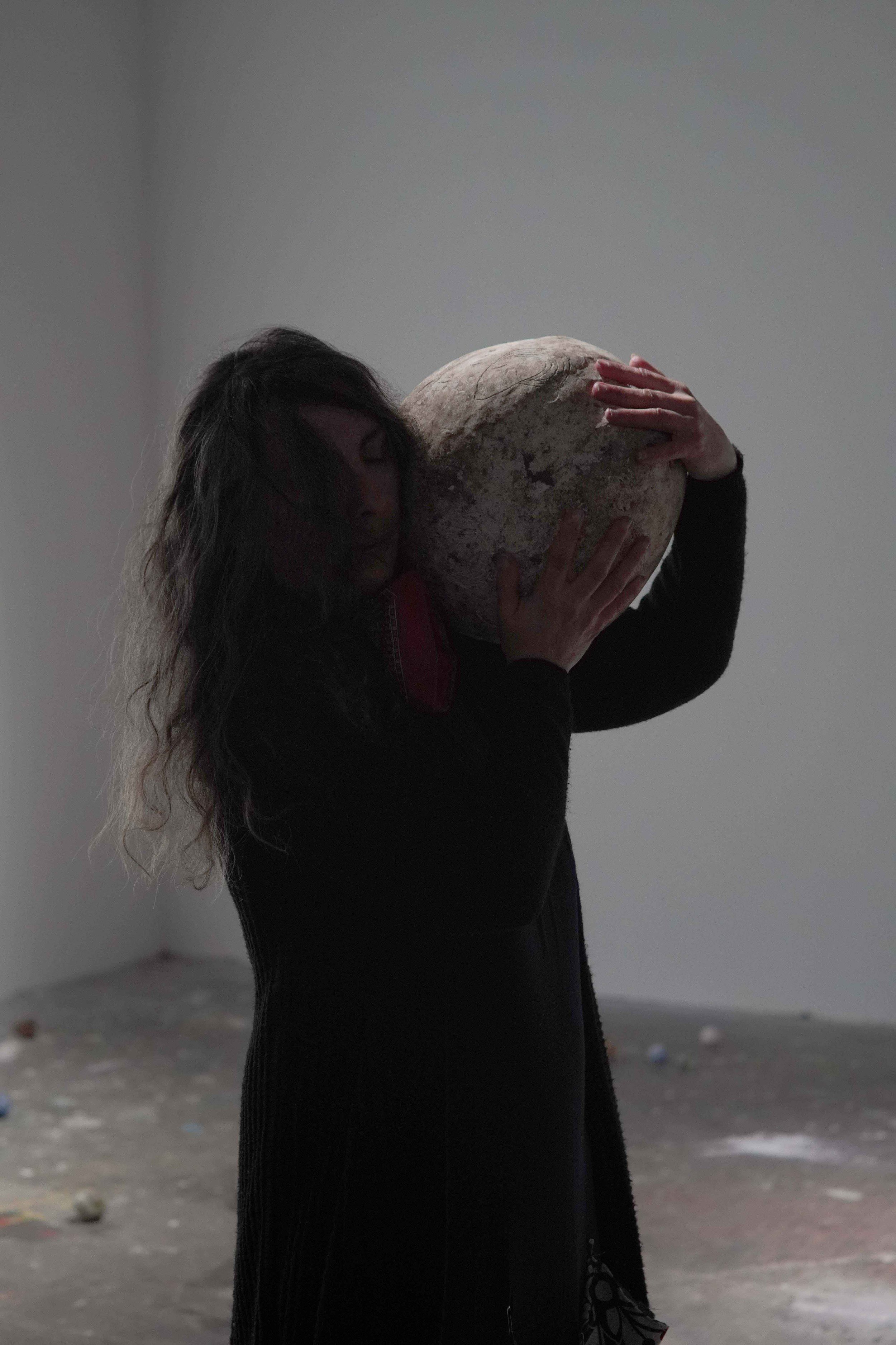 A woman with long curly hair facing away, holding a large round object, standing in a dimly lit outdoor area with a gray ground and scattered trash.