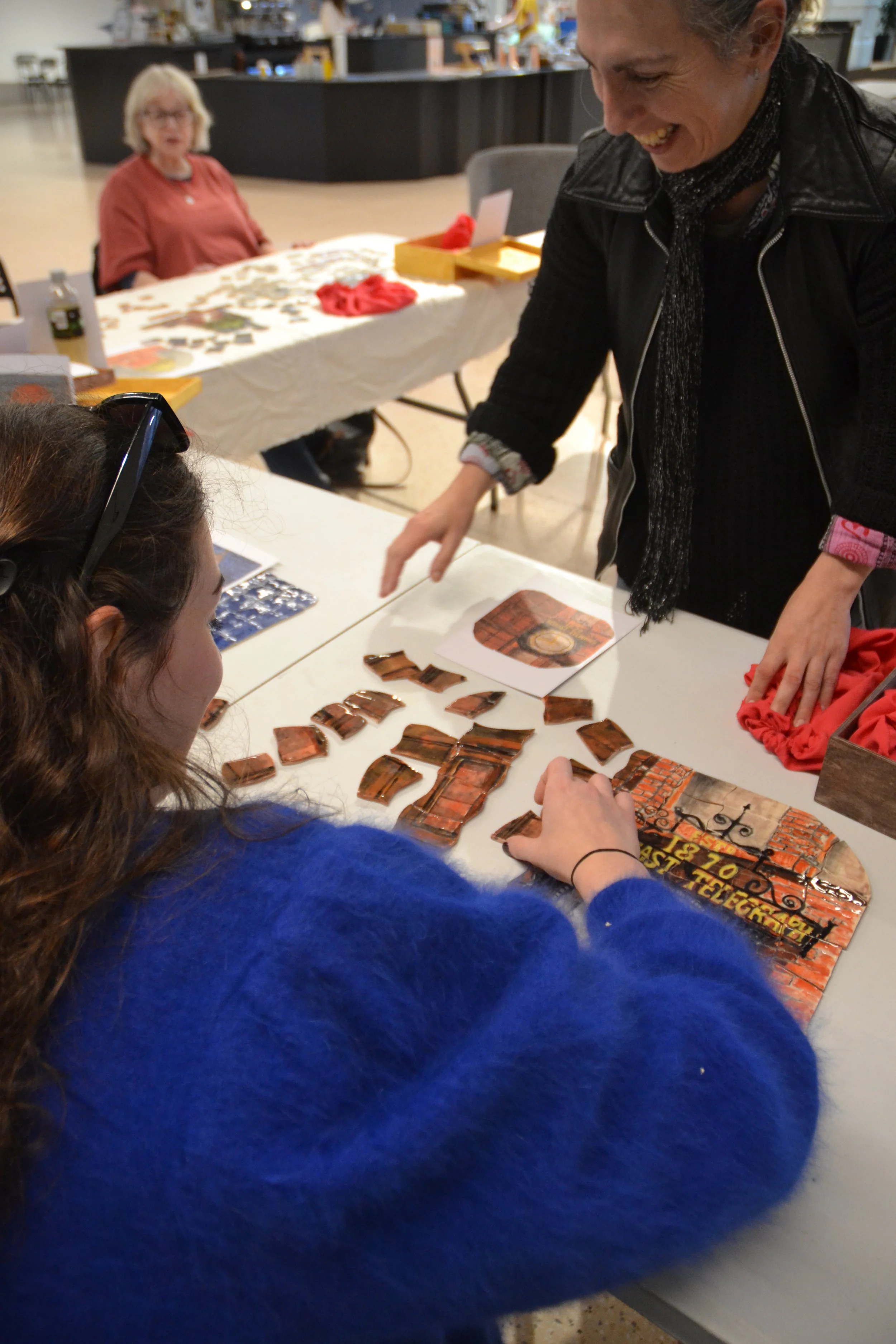 People playing a puzzle game at a table, with a woman smiling and interacting with a person in a blue sweater, in a public indoor space.