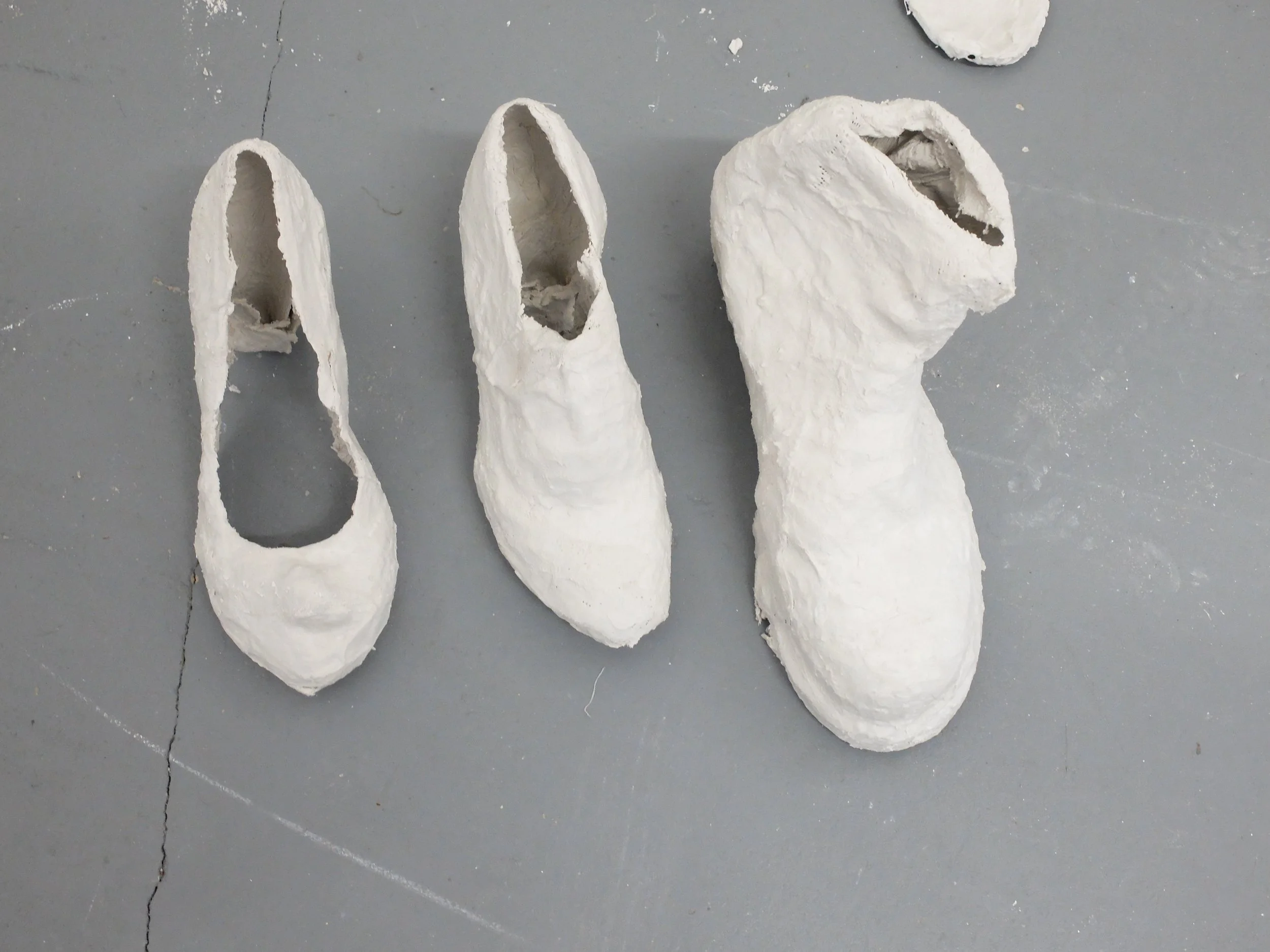 White ceramic or plaster molds of shoe shapes are placed on a gray floor, some with hollow interiors and openings at the top, used for casting or sculpting shoes.