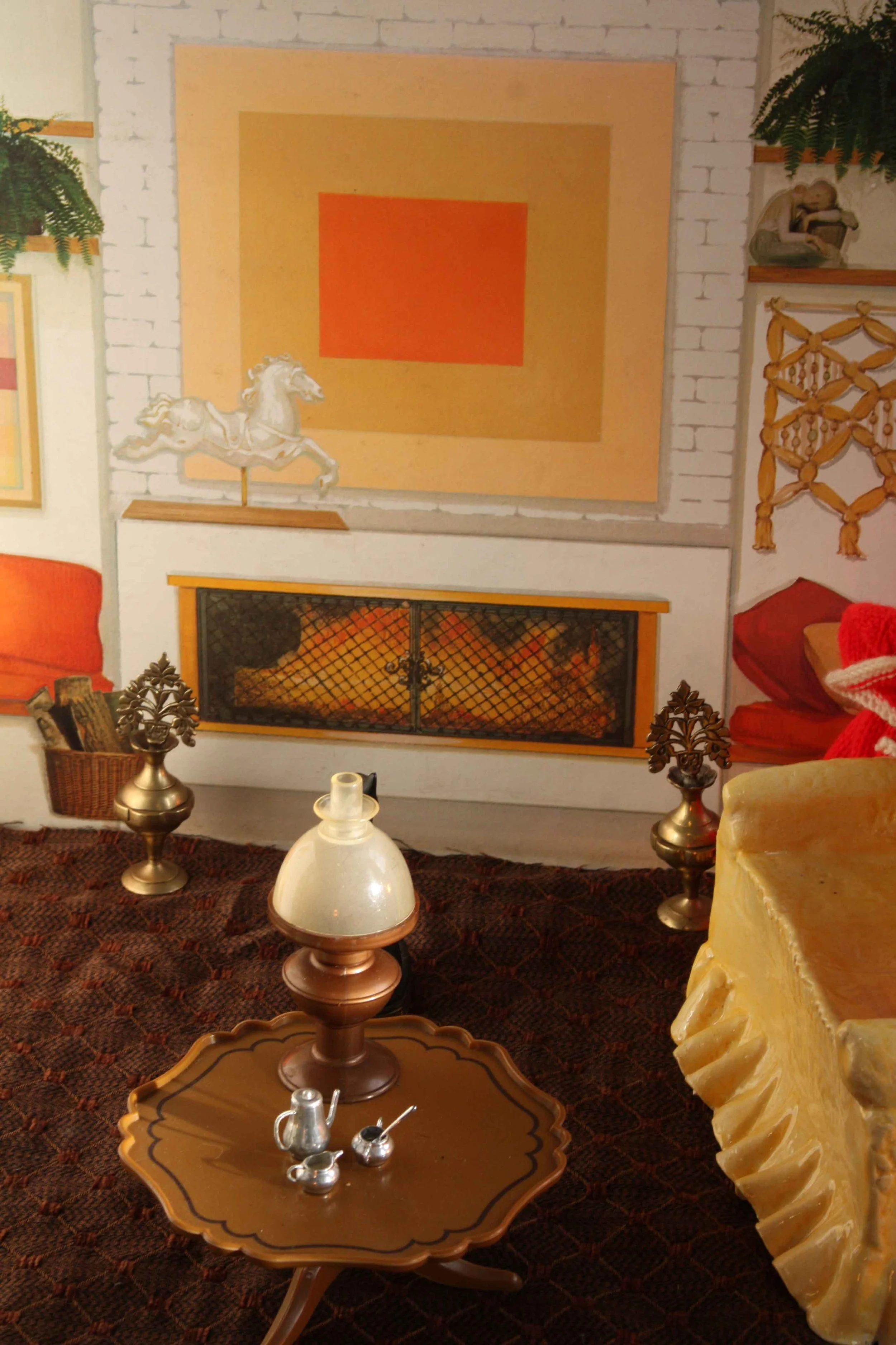 Interior toy  scene with a white brick fireplace, large abstract orange and beige painting on the wall, brass and wood decorative items, and a yellow armchair with red cushions on a rug.