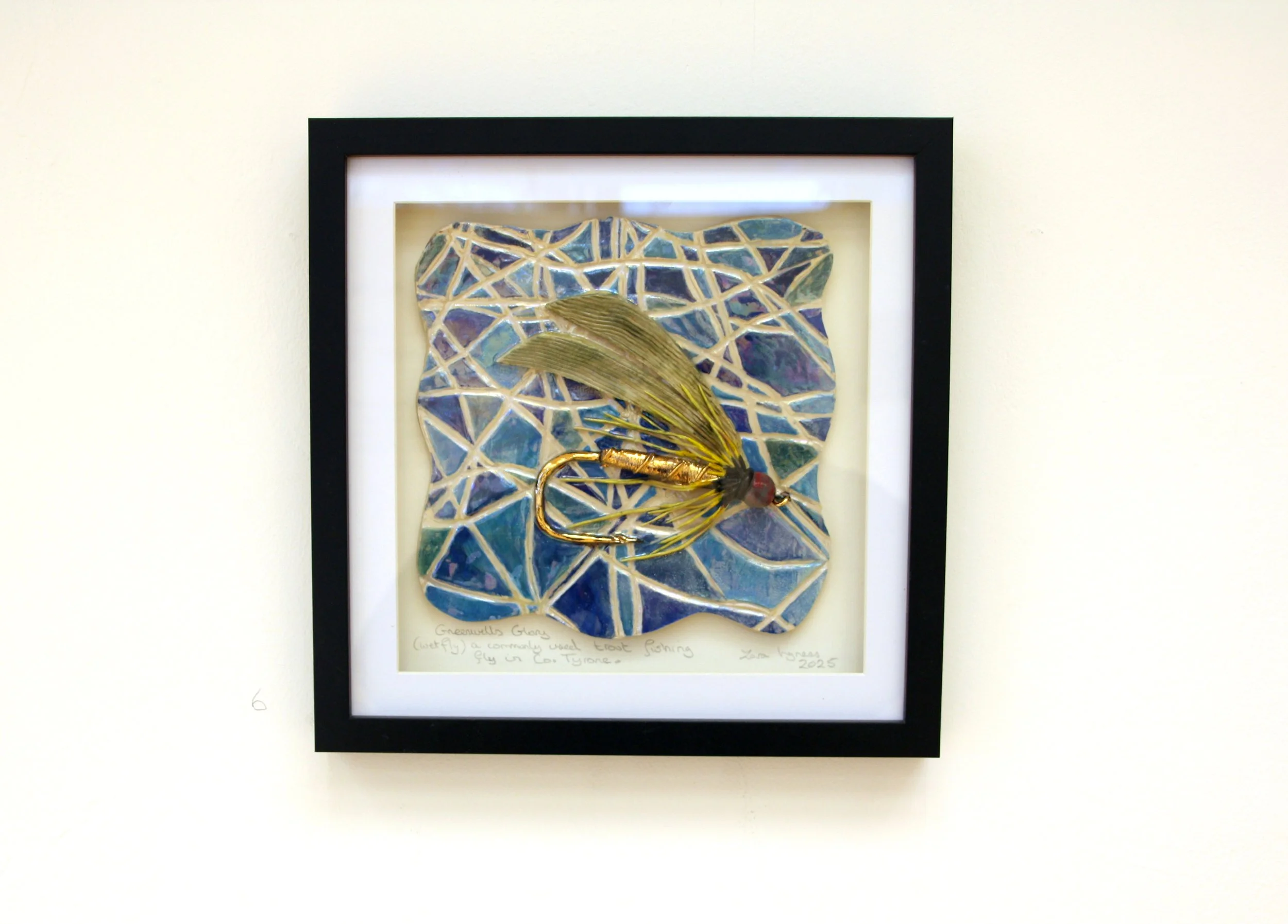 Framed artwork of a fishing lure with a mosaic background, displayed on a white wall.