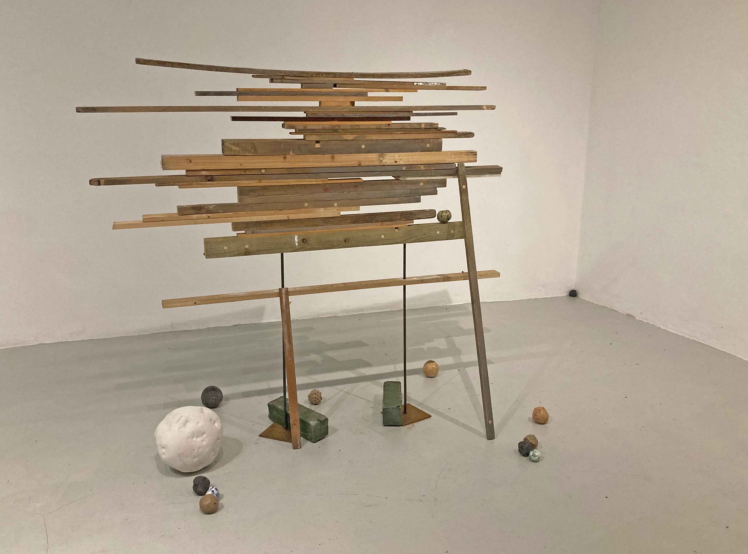 A sculpture composed of horizontal wooden planks arranged in a vertical form with various objects and textured balls at the base and around it, set against a plain white wall.