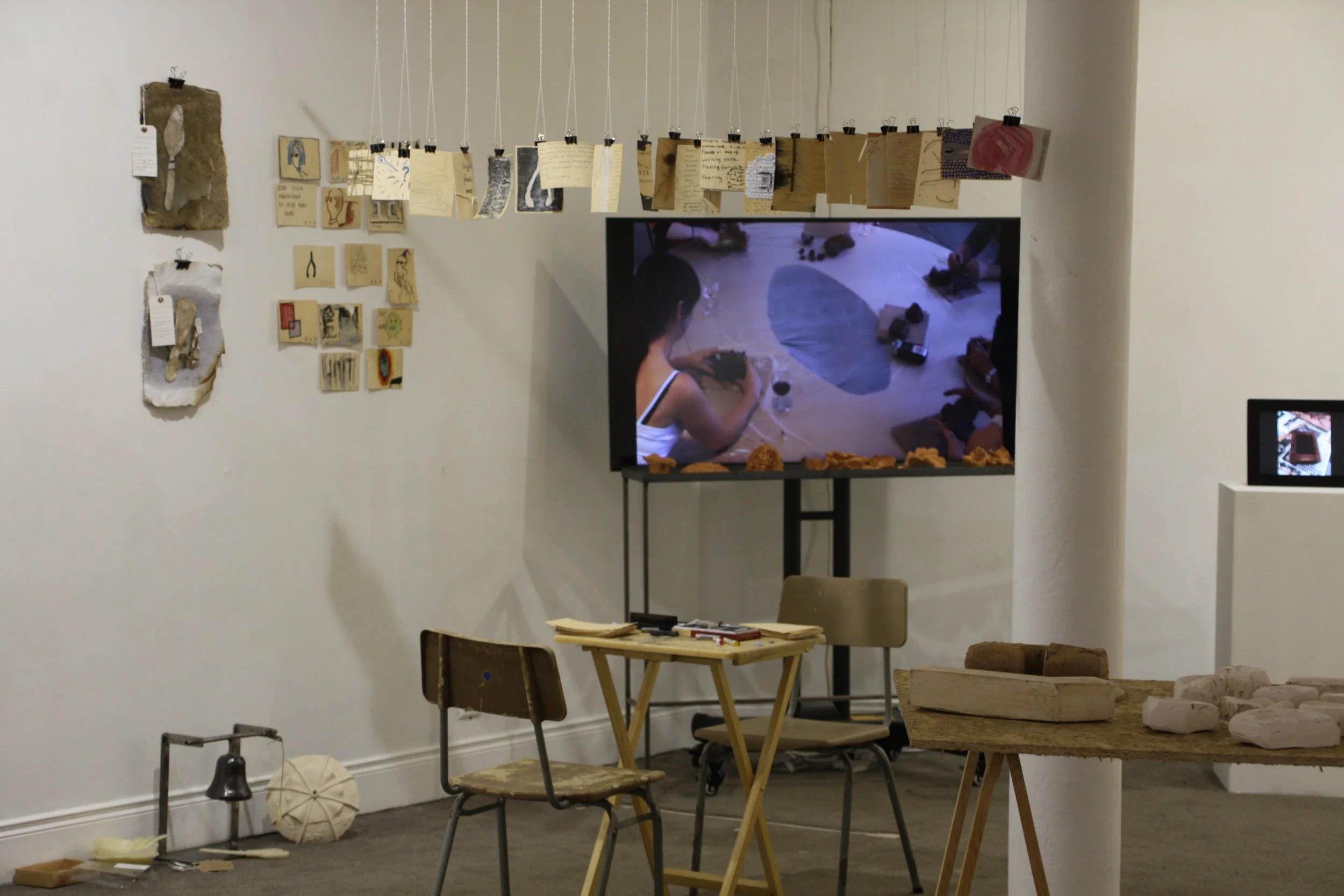 An art installation with hanging paper pieces, a television screen displaying a woman working on art, and various small objects and artworks arranged on tables and walls.