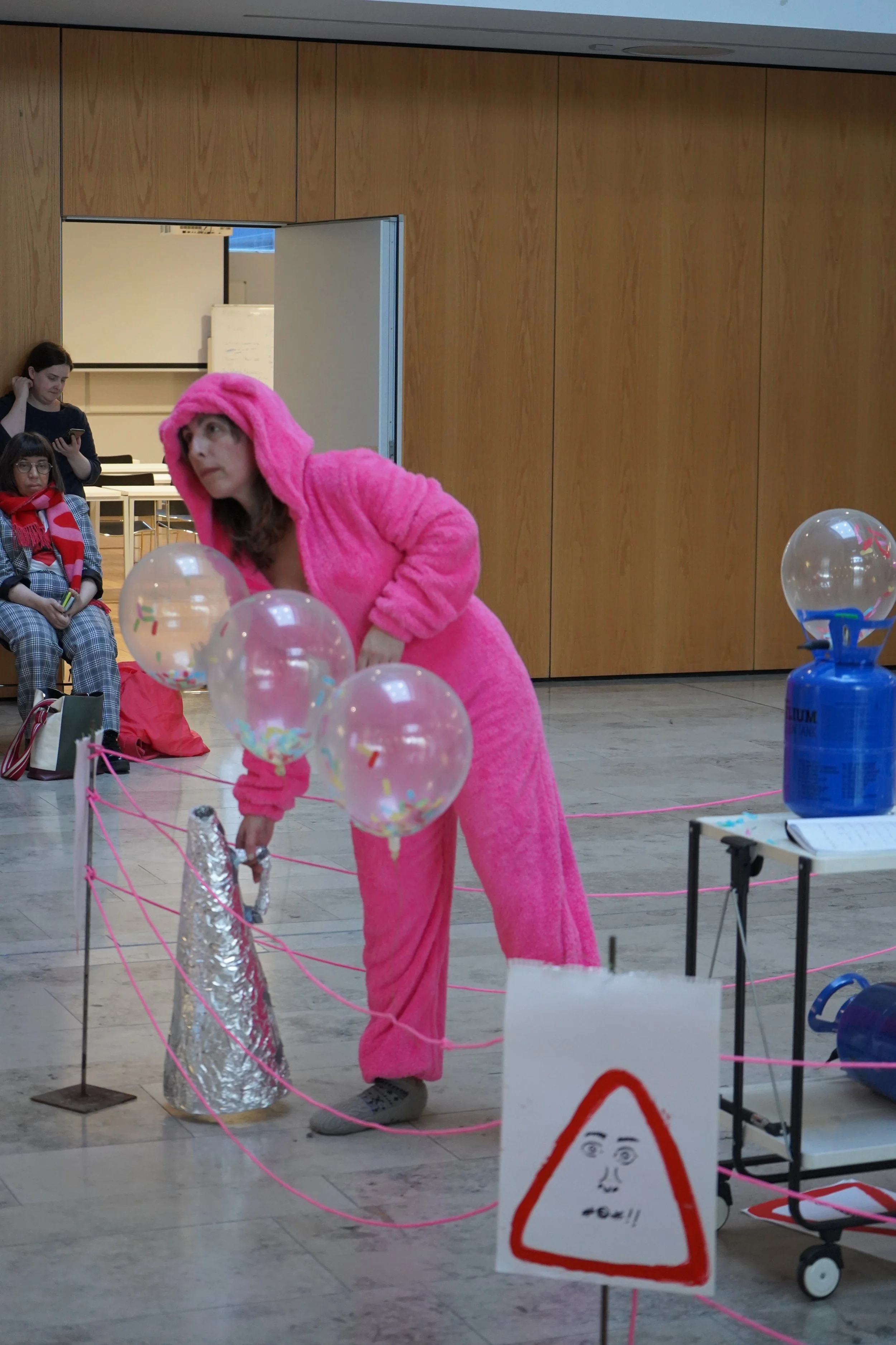 Person dressed in a pink fuzzy costume, possibly resembling an animal or character, bending down near transparent balloons with confetti inside. In the background, there are others seated and standing, with a wooden paneled wall and various objects i