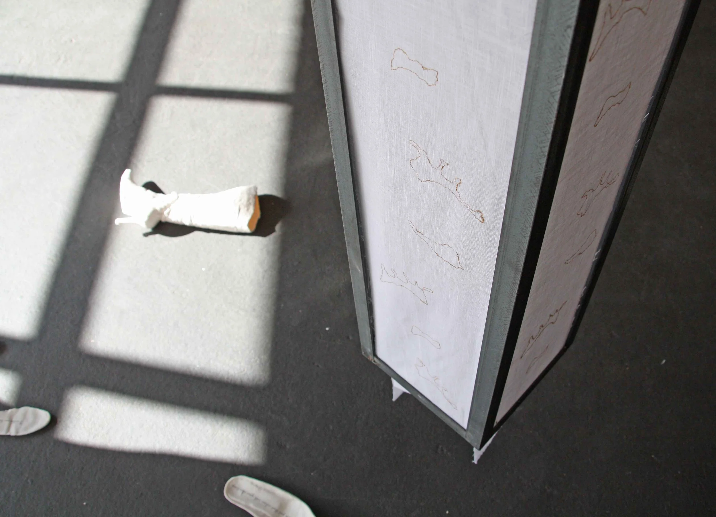 Close-up of an art display with a white canvas and thin gold outlines of abstract shapes, with a shadow cast on the backdrop of a metallic frame, and a small piece of white fabric or paper on the gray surface below.